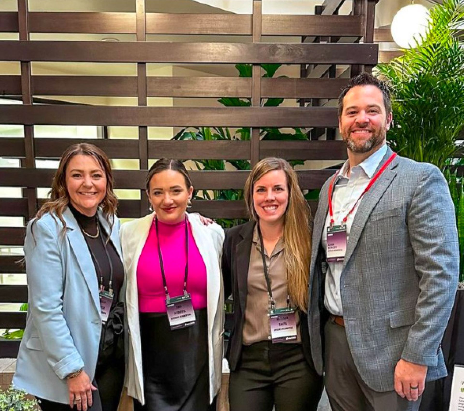 Four people posing indoors at a networking event, wearing conference badges, smiling in front of a wood slat wall.