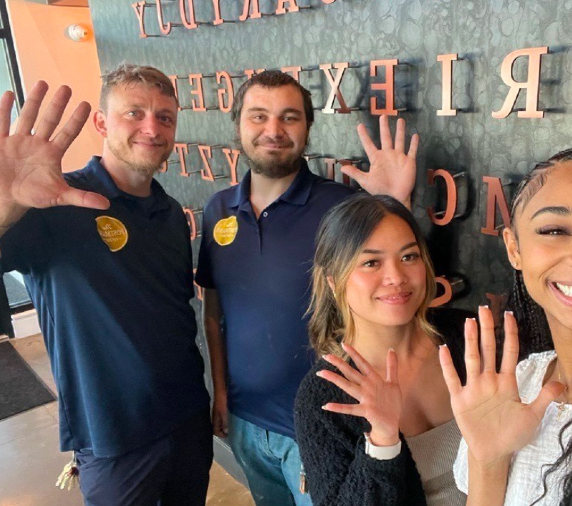 Four people smiling and waving indoors in front of a wall with mirrored lettering