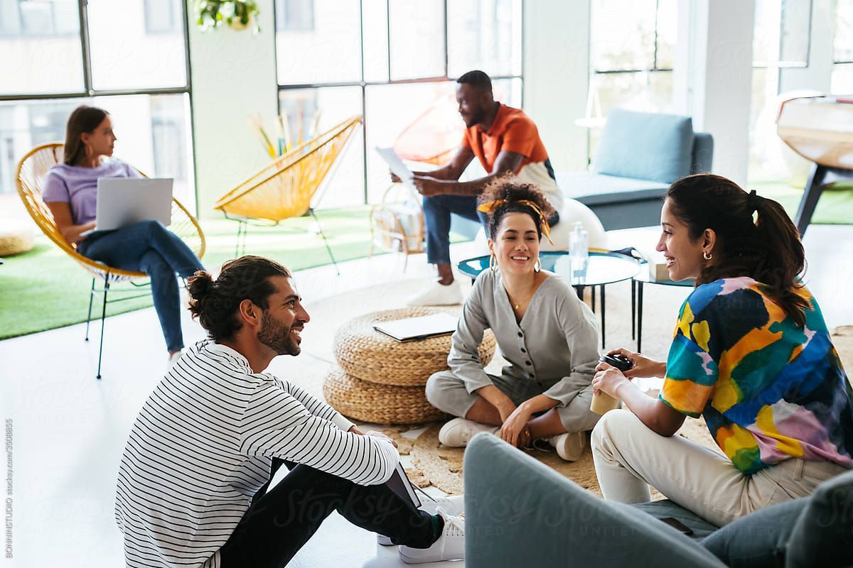 People chatting in a bright living room, seated on couches and chairs with laptops and coffee.