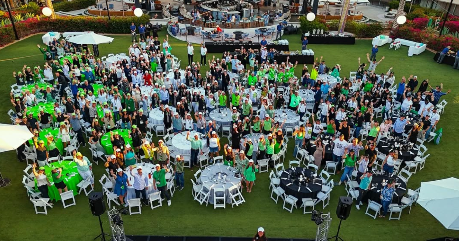 Large outdoor gathering with many people seated at round tables on a lawn, viewed from above.
