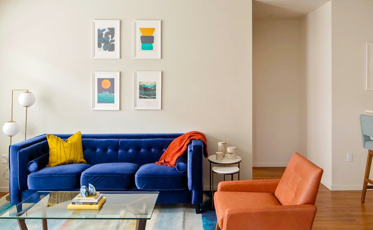 Bright living room with a blue sofa, orange chair, glass coffee table, and framed wall art.