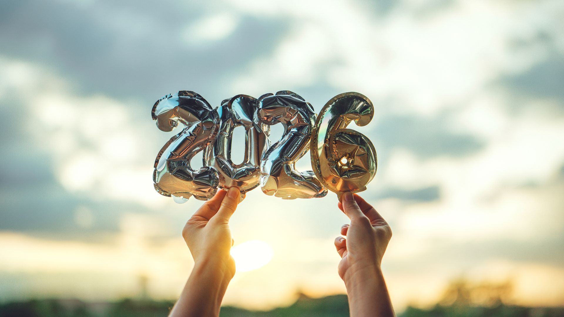 Hands holding metallic balloon numbers “2026” against a sunset sky