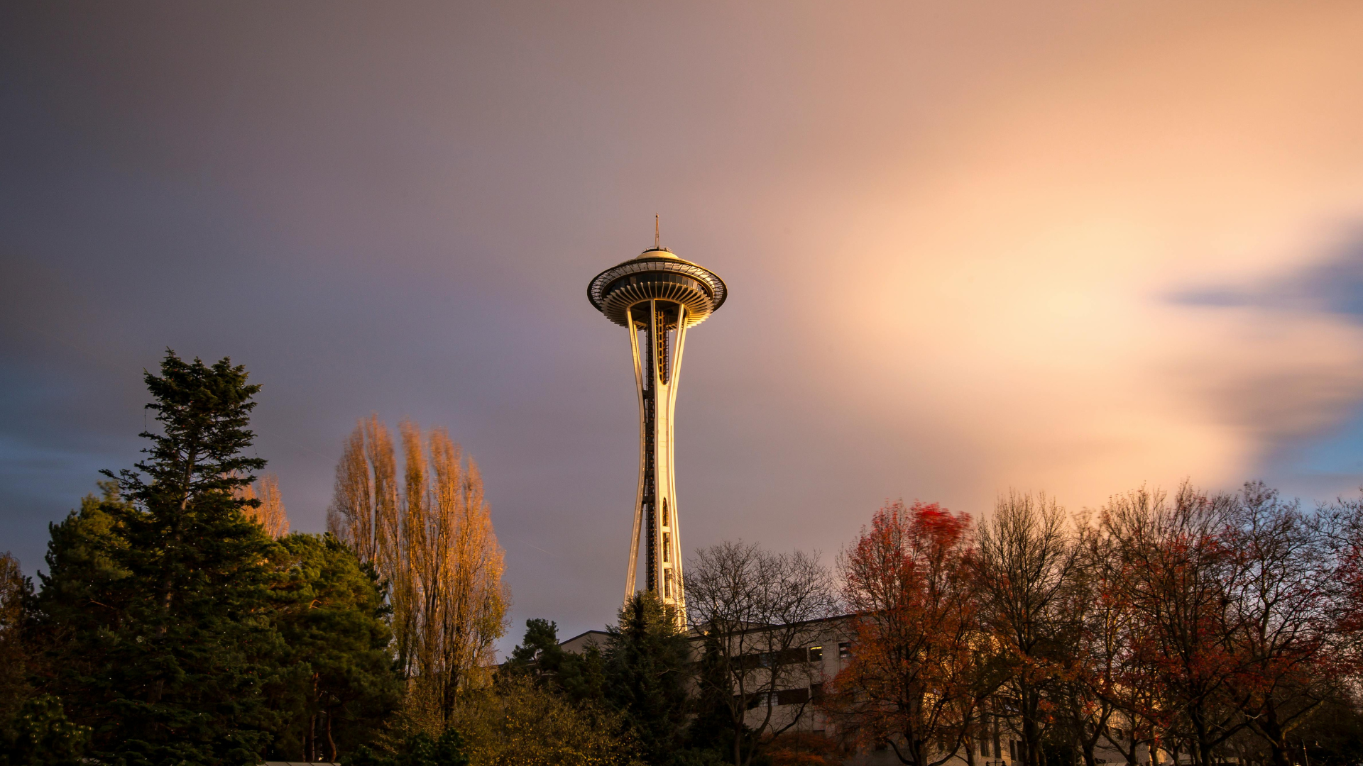 Space Needle above autumn trees at sunset, with a glowing sky and clouds behind it