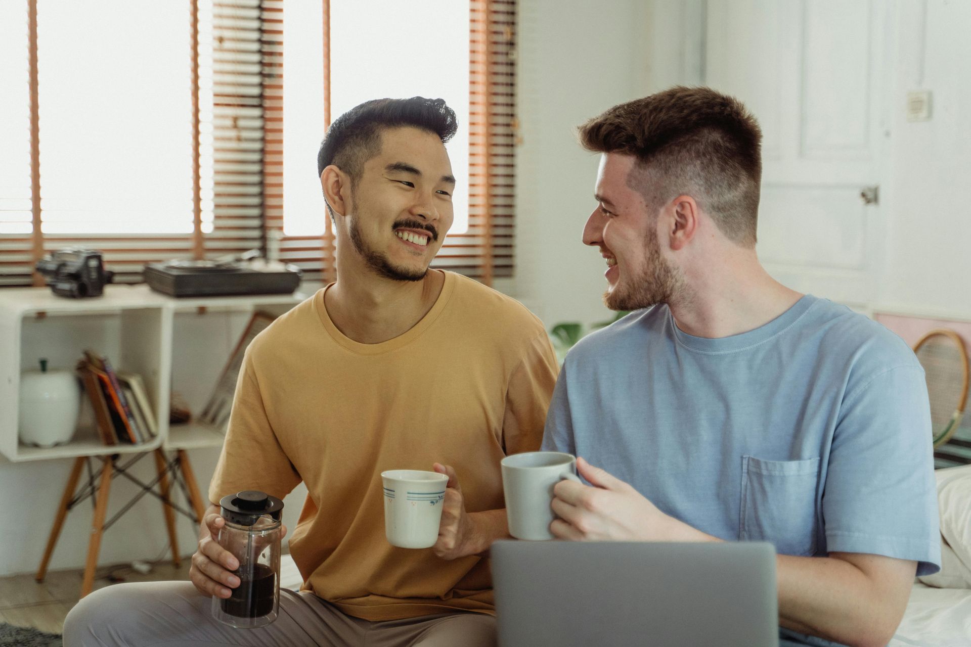 Two people smiling and chatting with coffee mugs beside a laptop in a bright home office