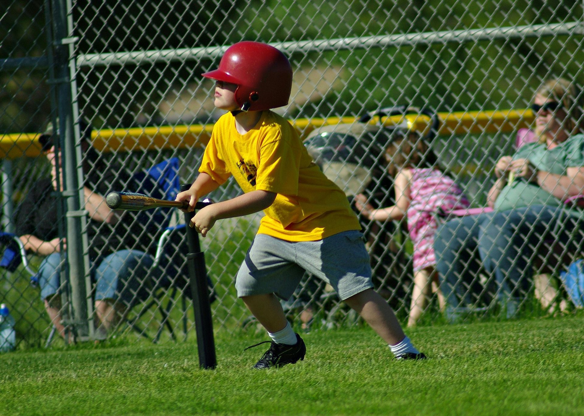Child in red helmet and yellow jersey running on a baseball field with spectators behind a chain-link fence