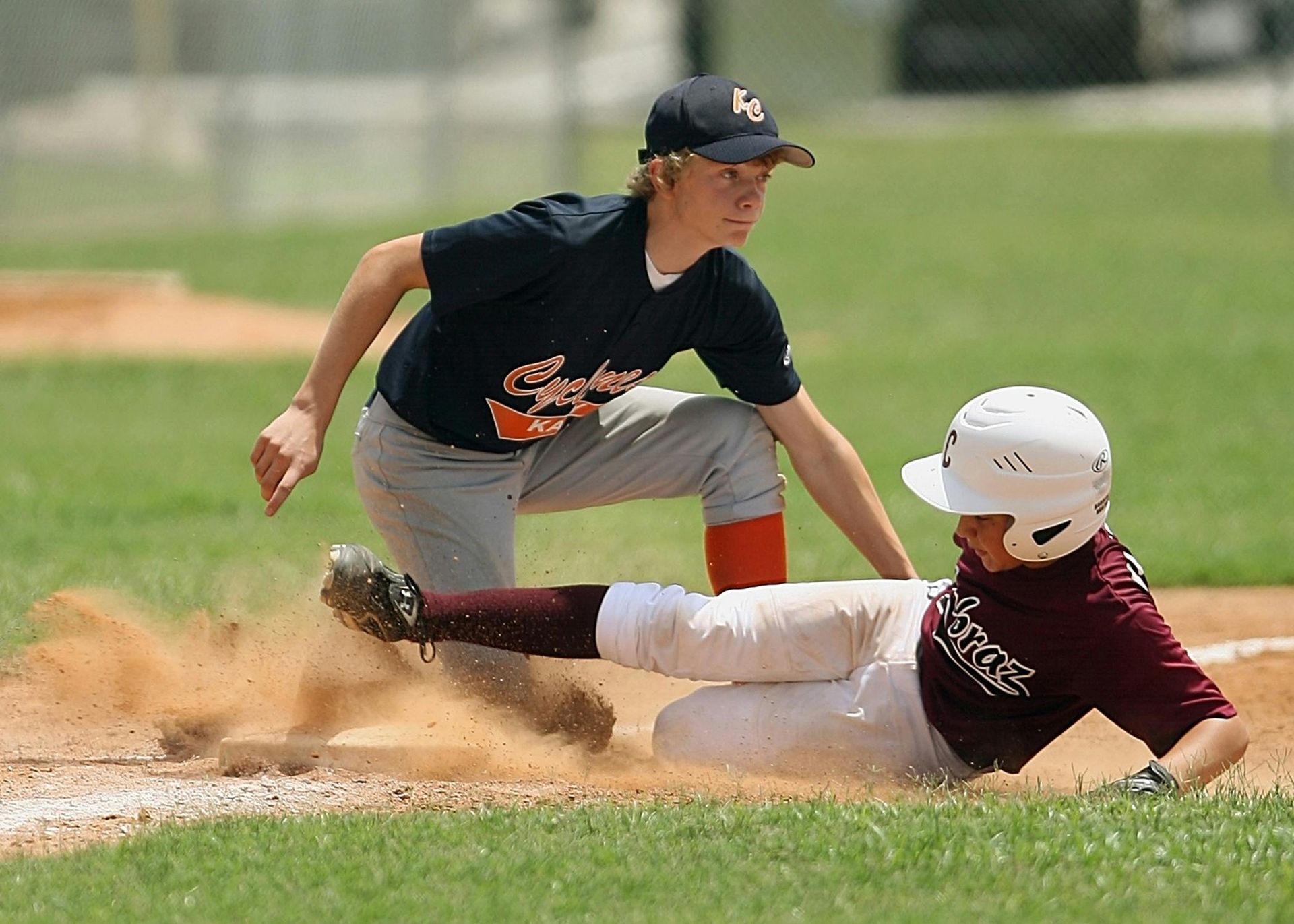 Baseball player sliding into a base as a fielder reaches to tag him, kicking up dust.