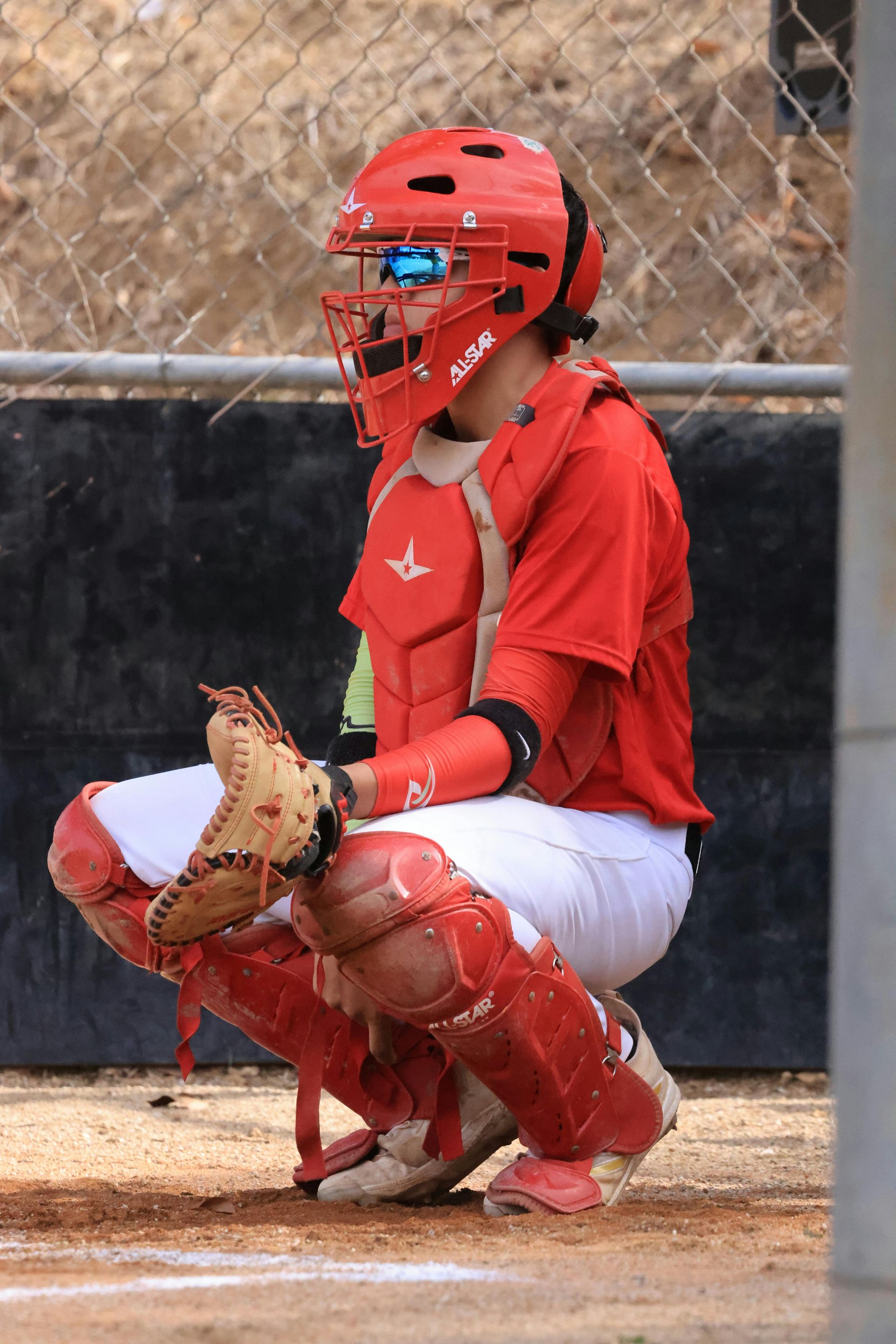 Red softball catcher in full gear crouching behind home plate, holding a glove on the field.