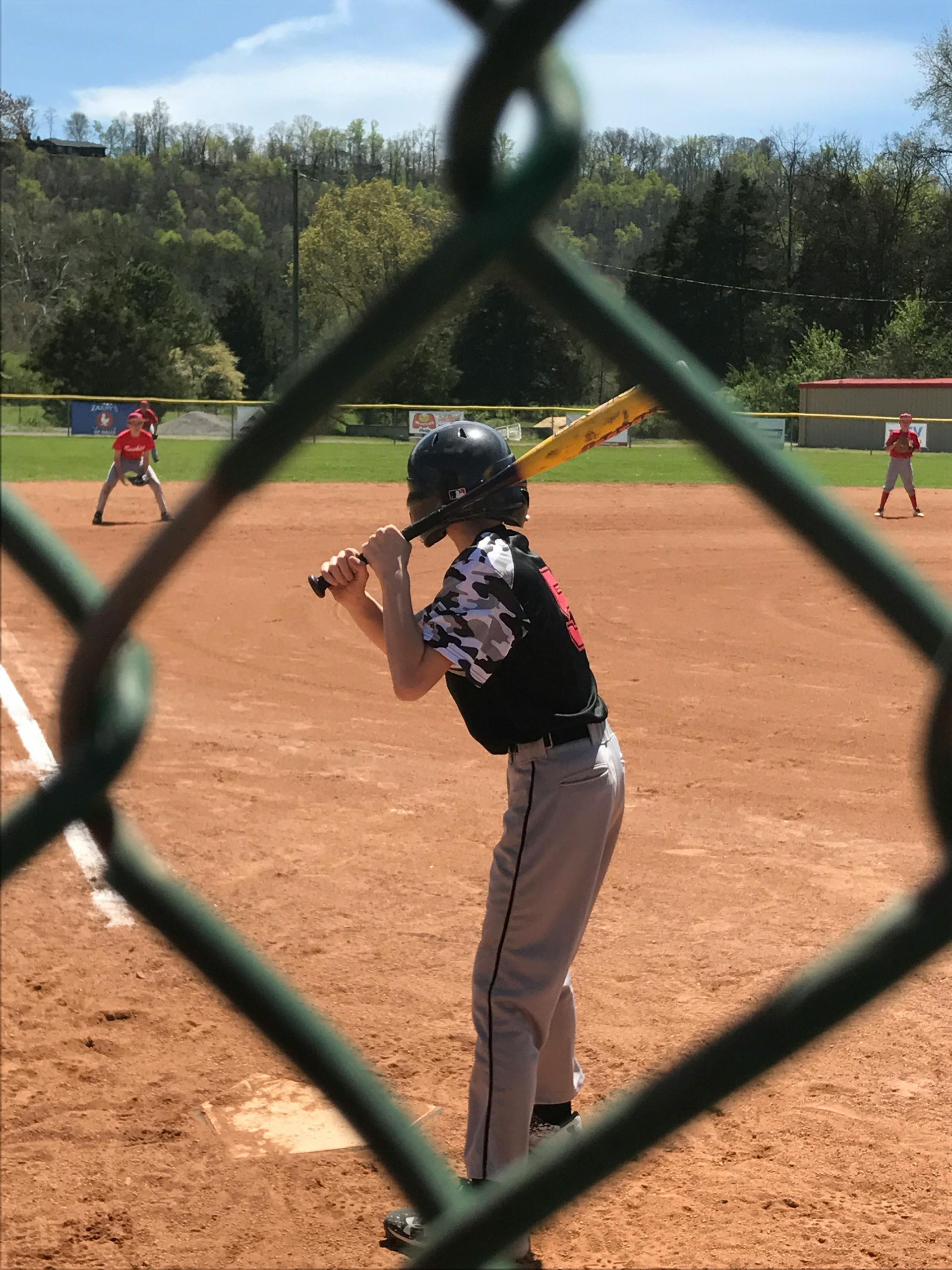 Baseball batter at the plate behind a chain-link fence, with fielders in the background.