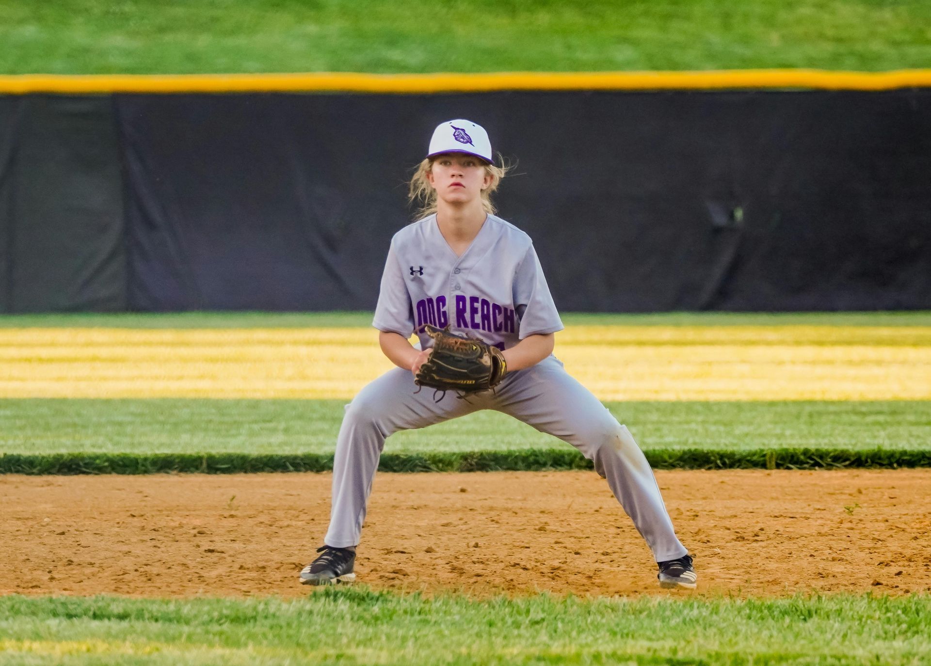 Youth baseball player crouches on dirt field, glove ready, in gray uniform and cap.