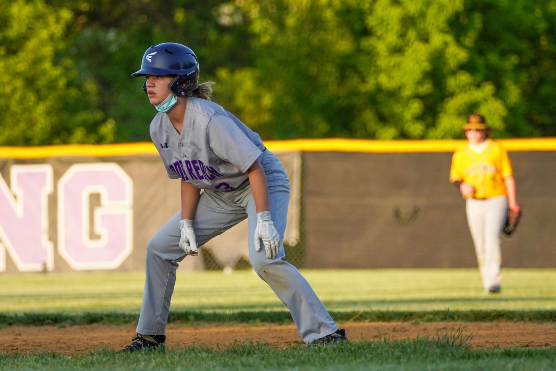 Softball player in gray uniform and blue helmet ready on the field, with an outfielder in the background.