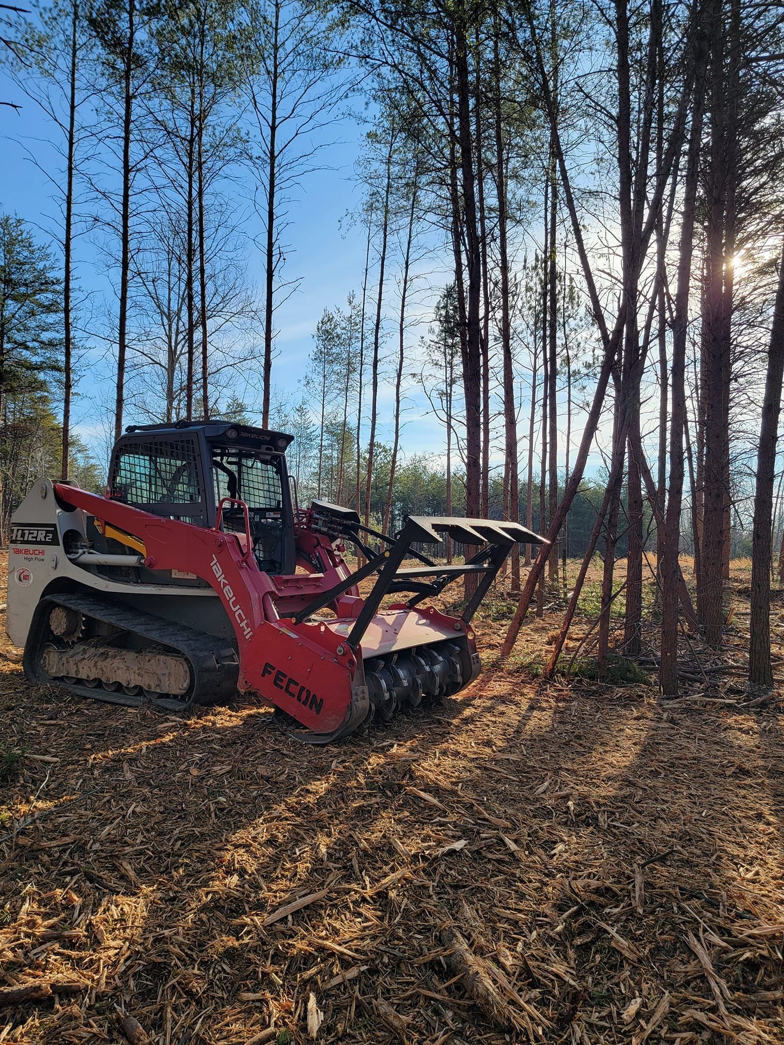 A red and white Takeuchi track loader with a Fecon forestry mulcher attachment in a forest clearing.