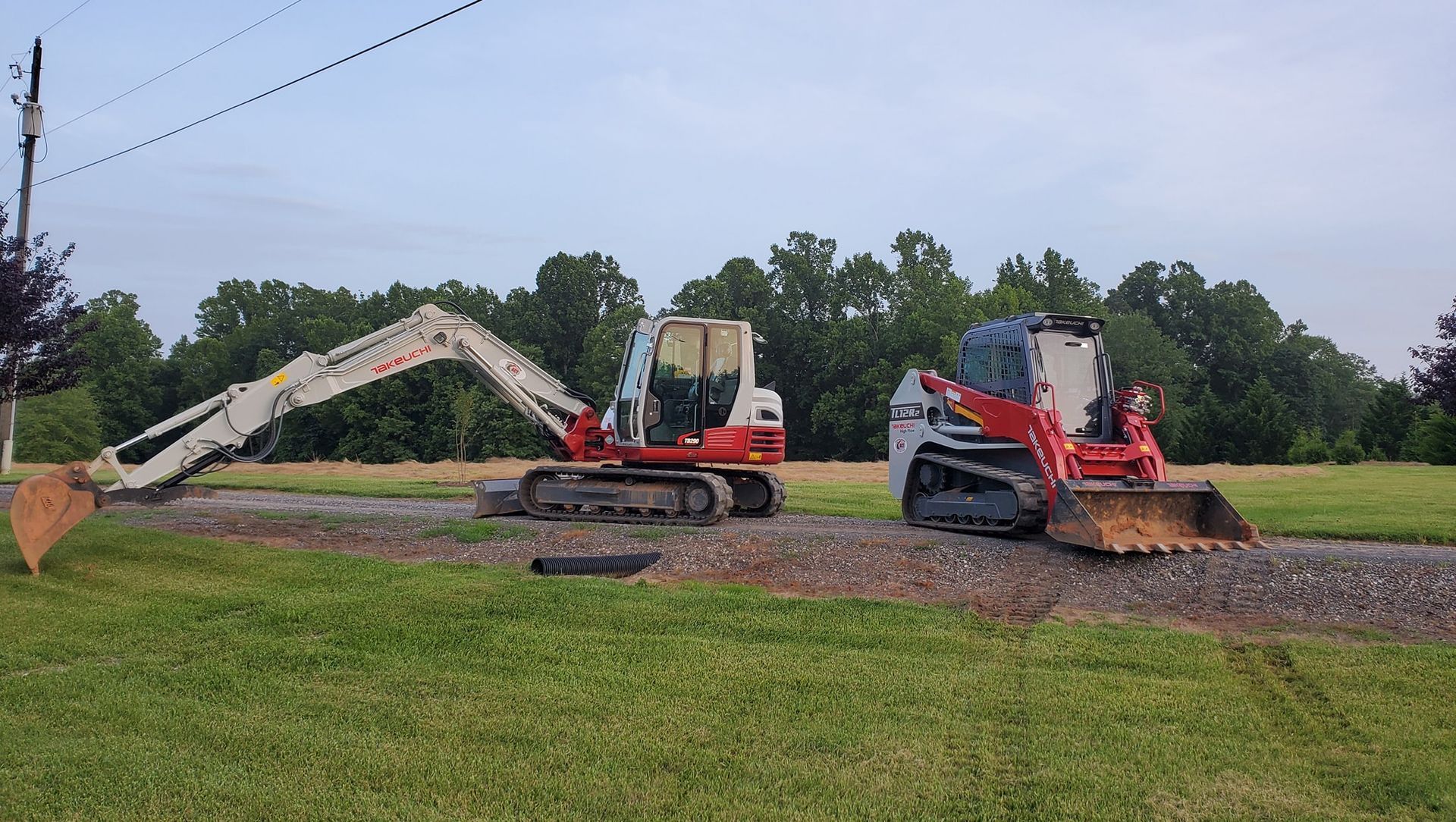 A white excavator and a red tracked skid steer parked on a gravel path in a grassy field under a cloudy sky.
