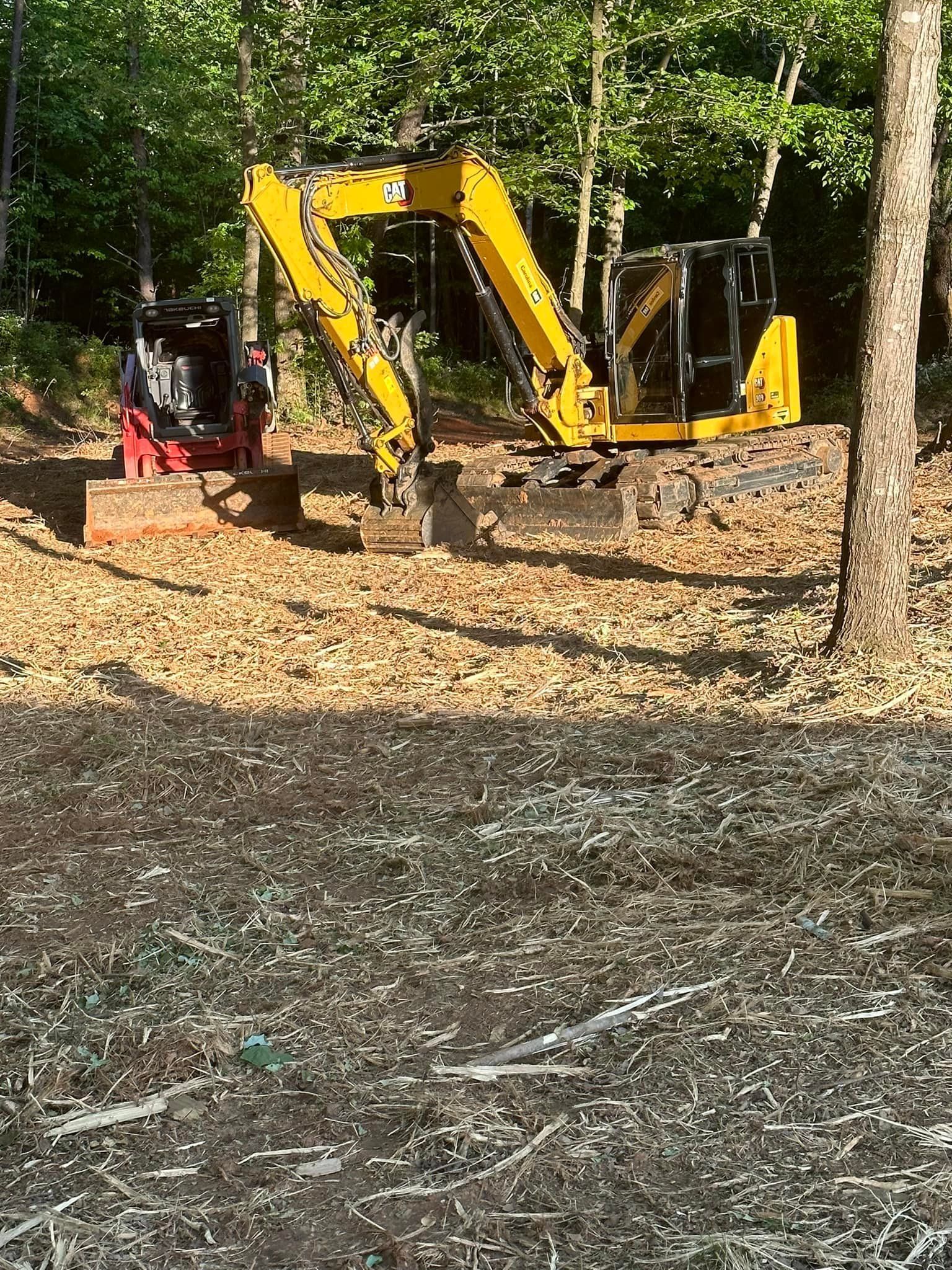 A yellow excavator and a red skid steer parked in a wooded area on a dirt clearing covered in fallen leaves.