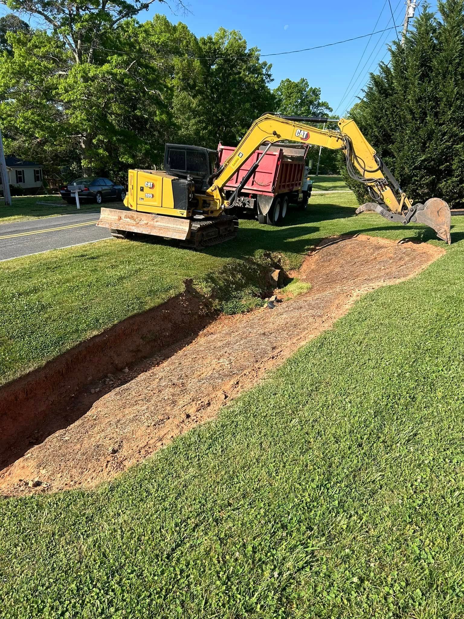 A yellow excavator sits in a grassy yard beside a long, freshly dug trench, with a red dump truck parked behind it.