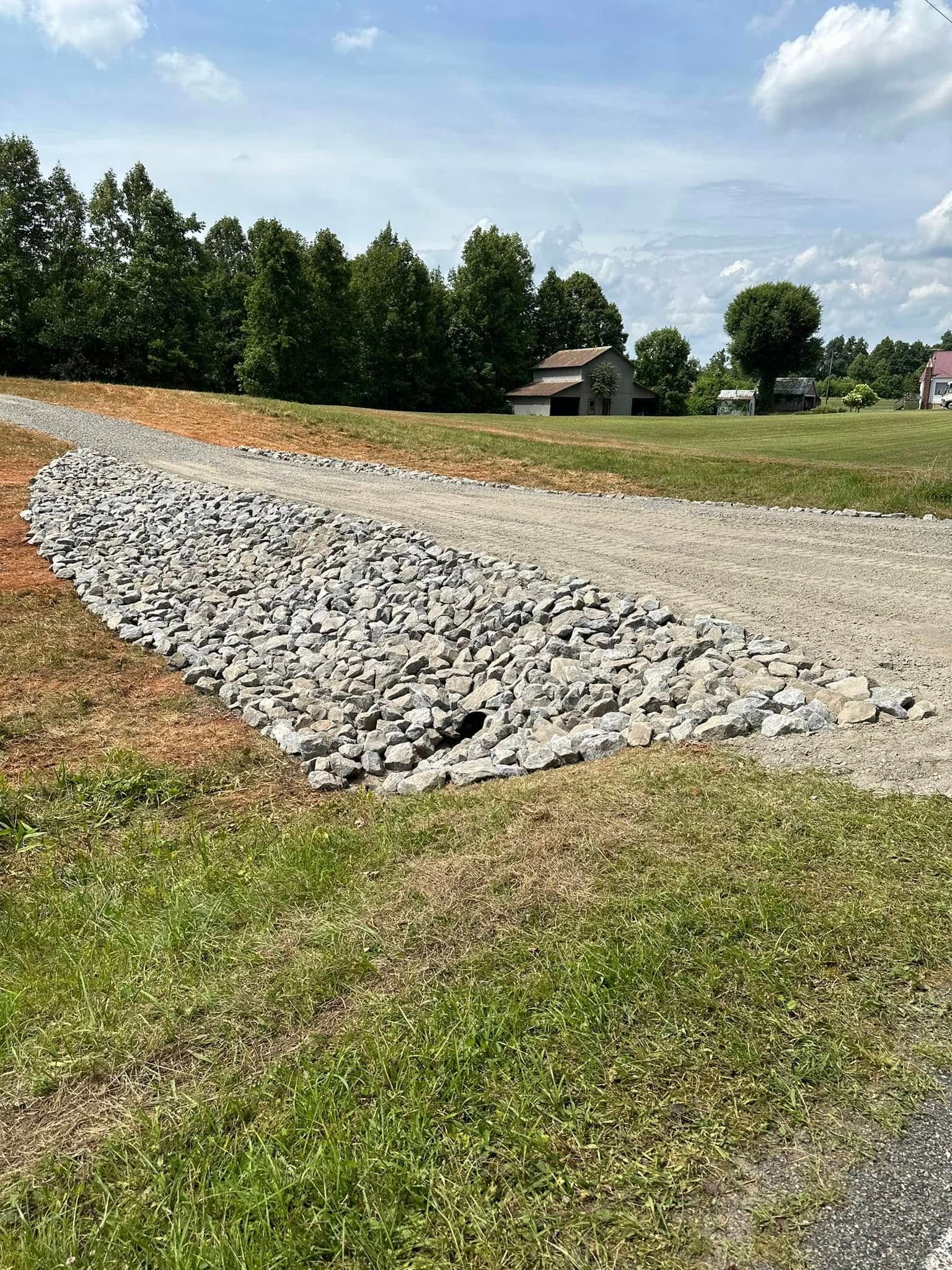 A drainage area filled with large grey rocks, situated alongside a gravel driveway in a grassy rural landscape.