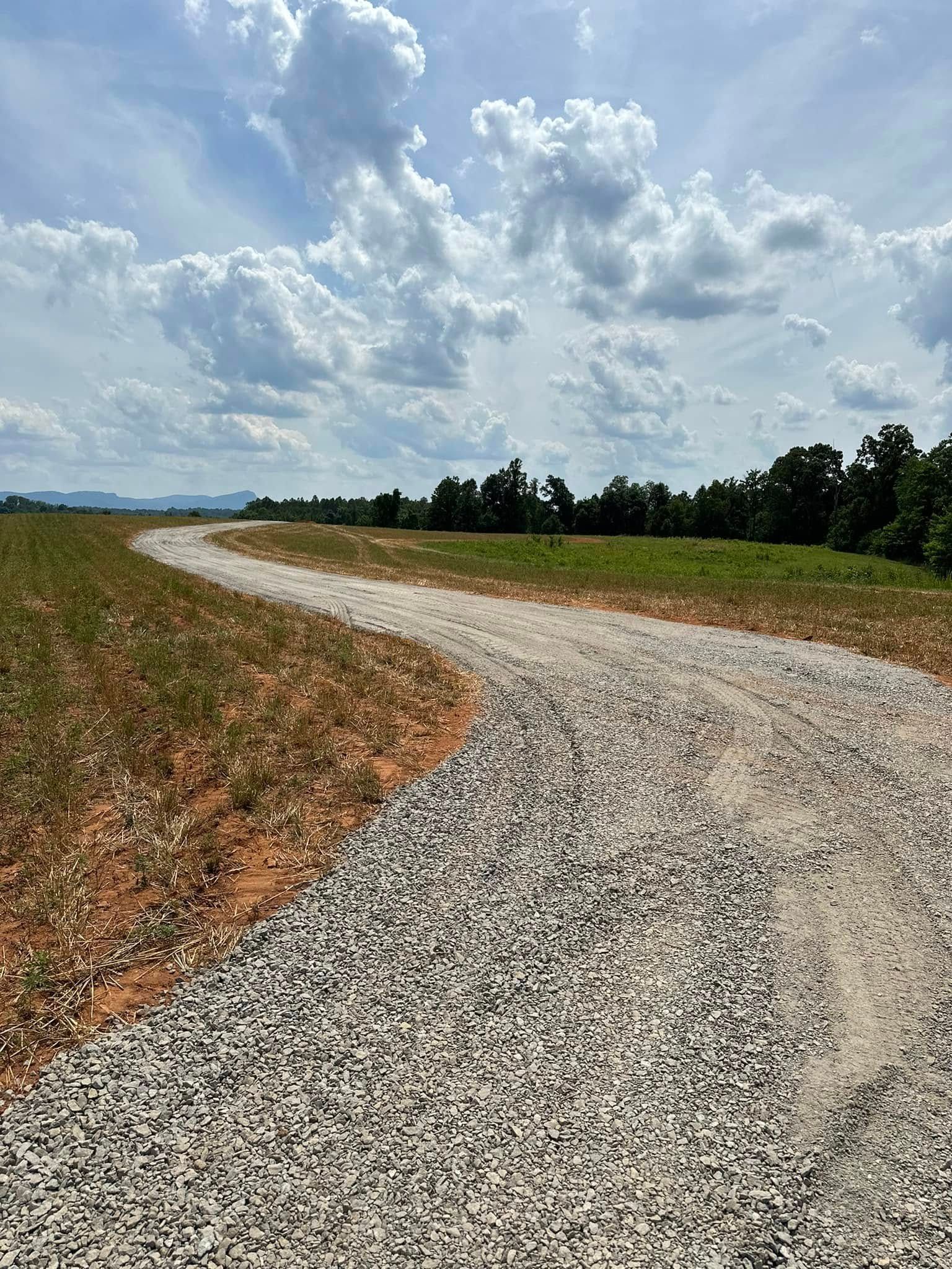 A gravel path curves through a field of dry grass and dirt toward a distant line of trees under a cloudy blue sky.
