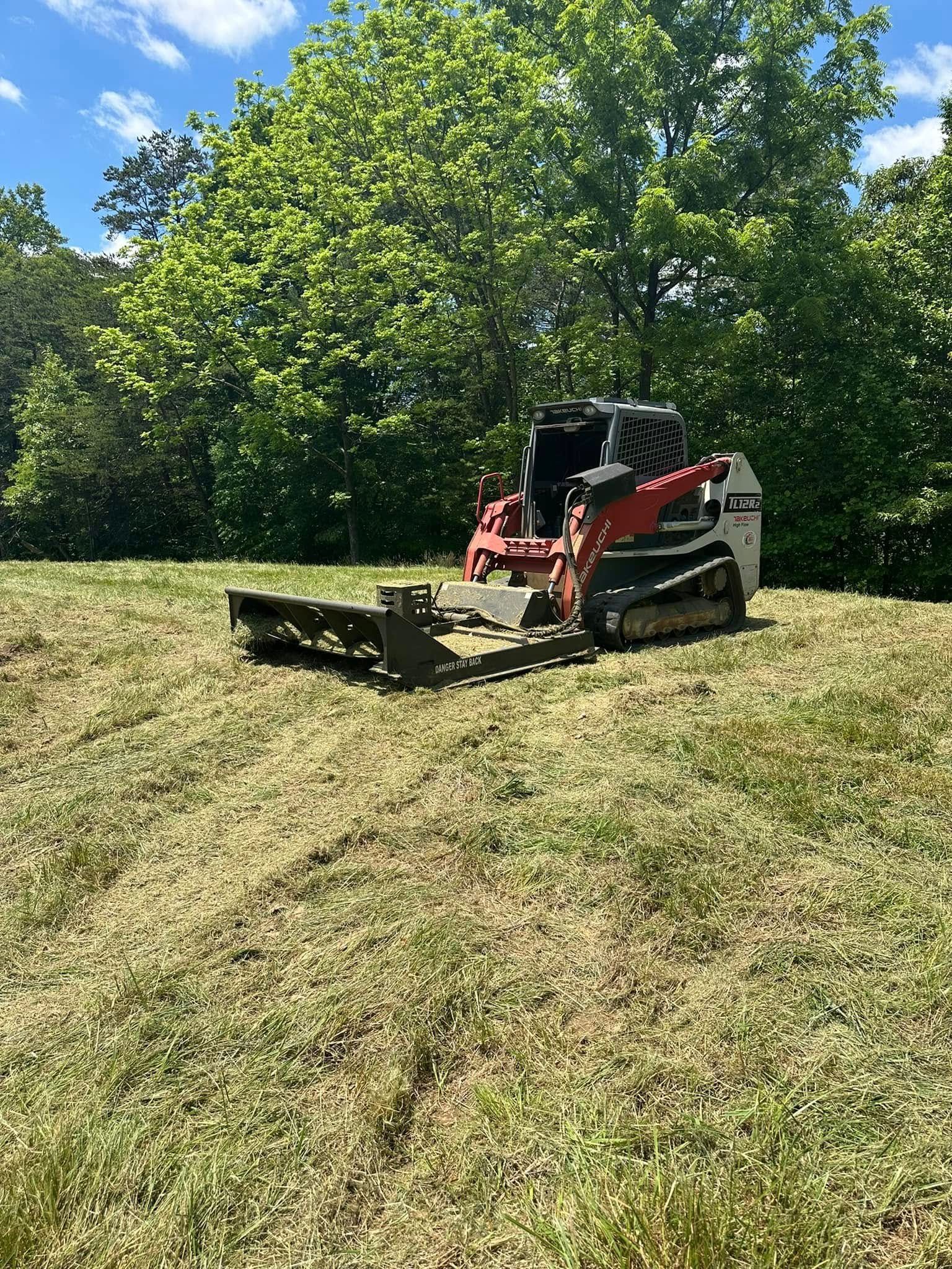 A red and white skid steer loader with a brush cutter attachment parked on a grassy hill in front of trees.