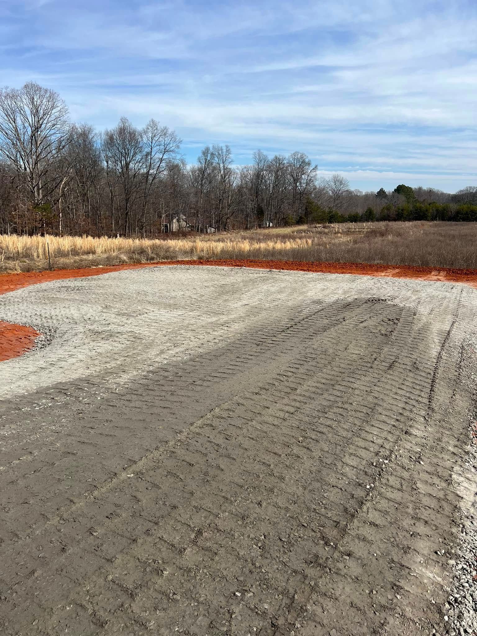 A gravel construction pad sits in an open field, with a line of trees in the background under a blue sky.