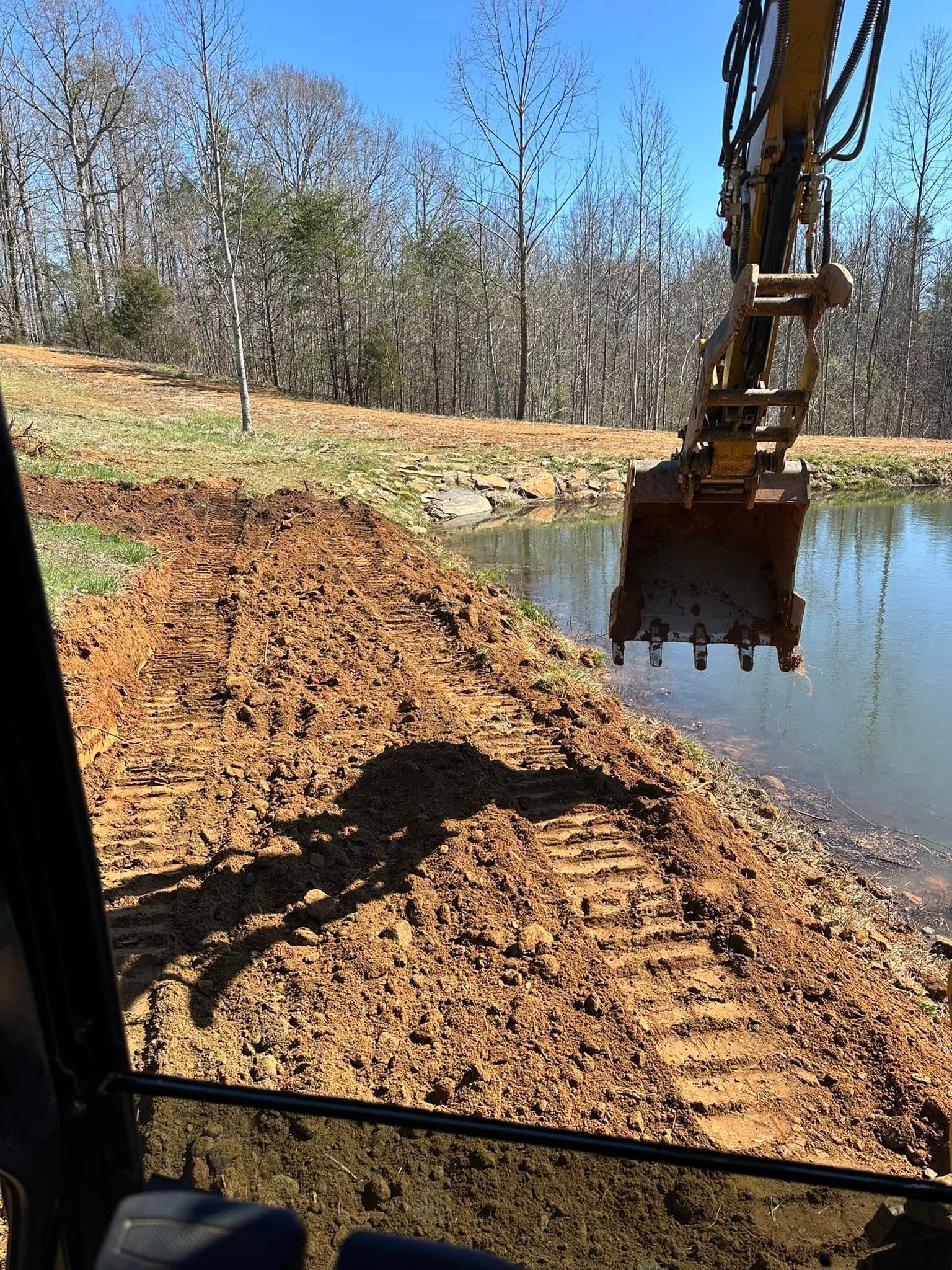 An excavator bucket suspended over a pond bank with freshly excavated soil, viewed from the operator's cab.