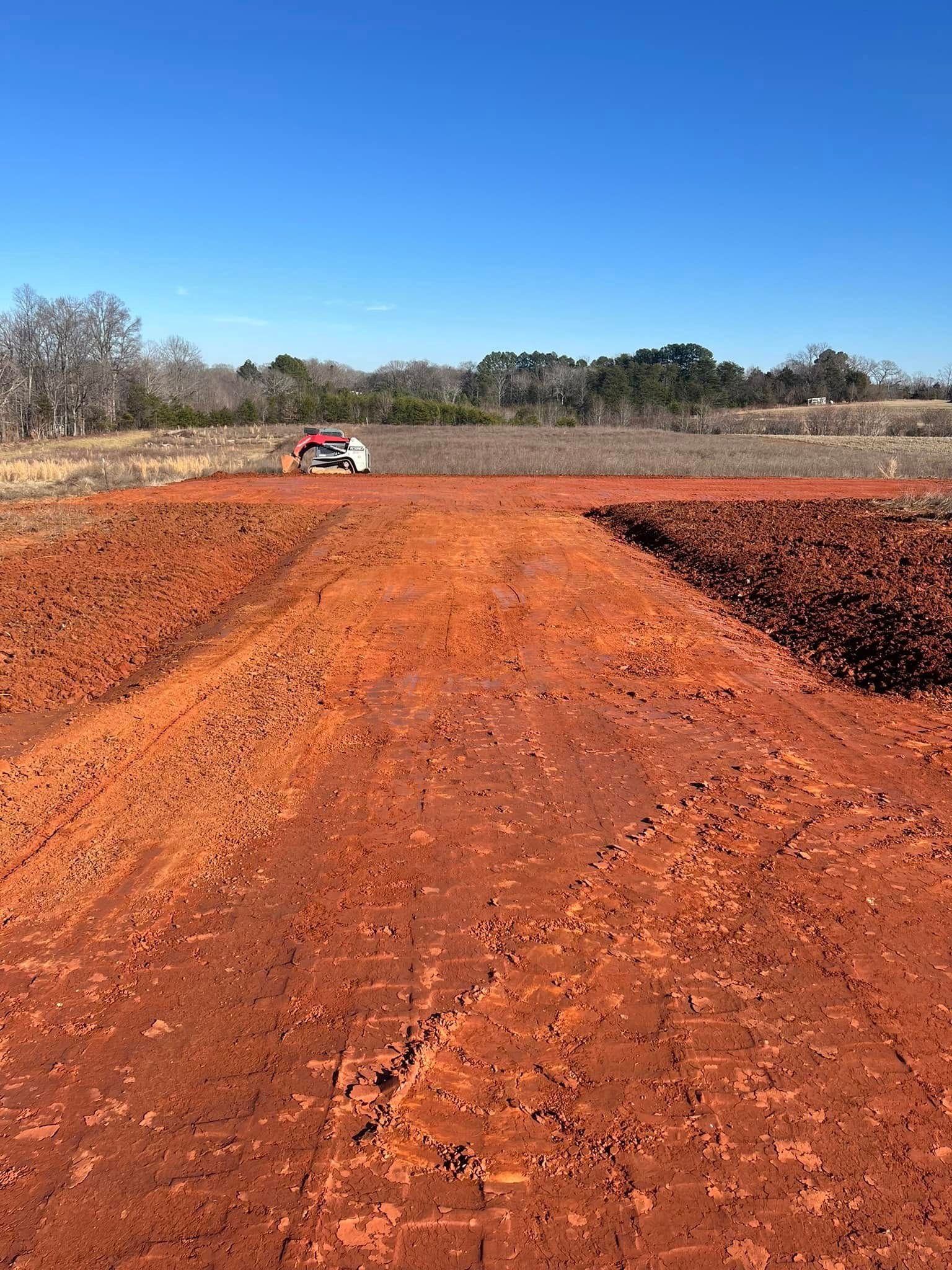 Construction equipment levels a plot of reddish-brown dirt under a clear blue sky in a rural setting.