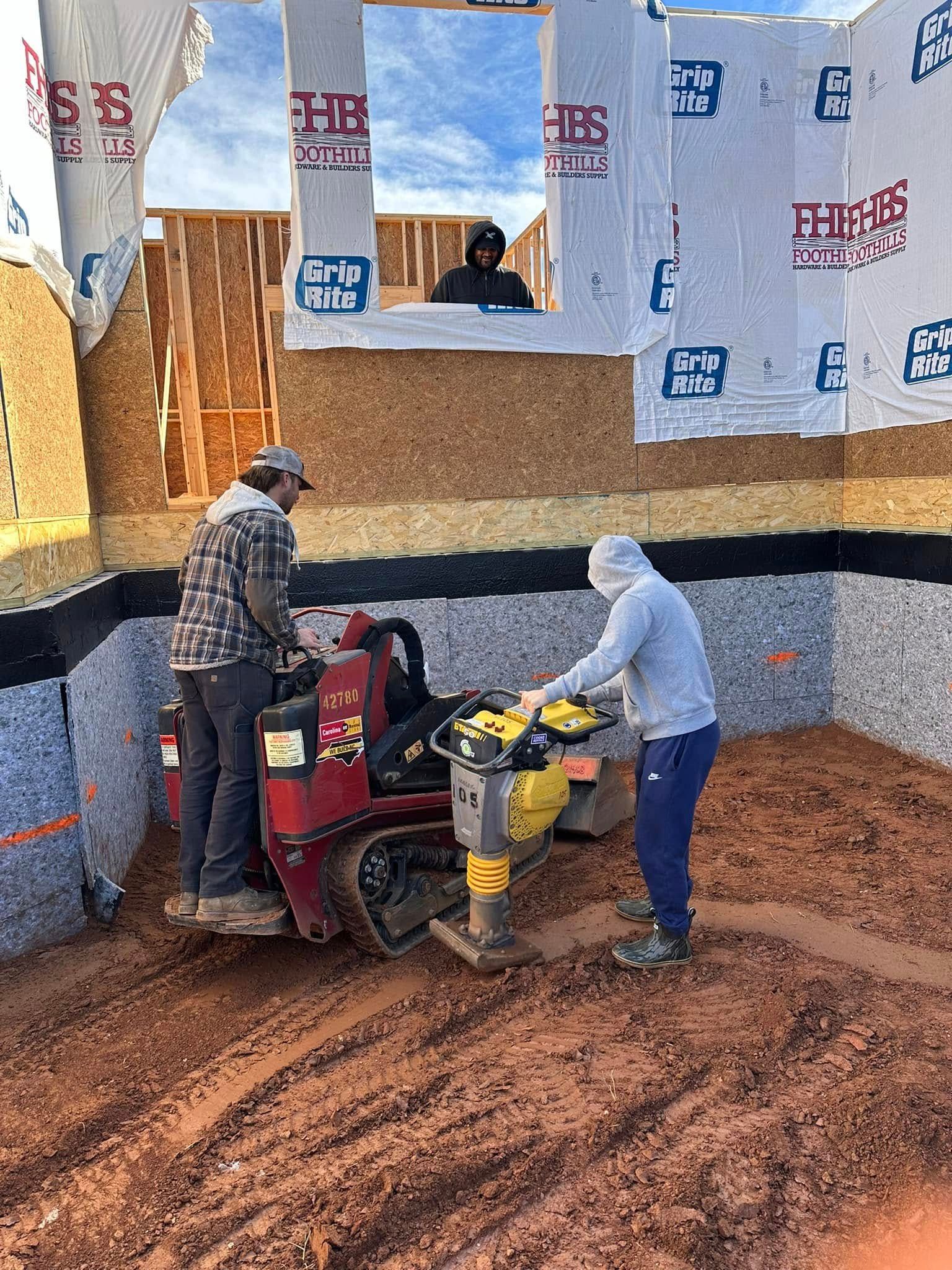 Three people work inside a partially built structure, using a walk-behind compactor on red dirt ground.