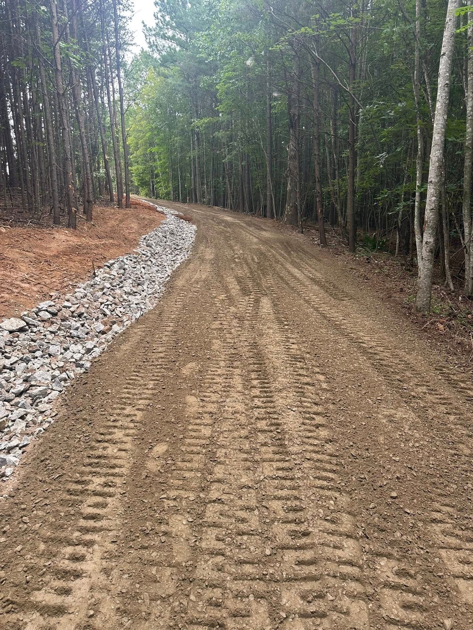 A dirt path with tire tracks leads through a forest, bordered by a drainage ditch filled with grey rocks on the left.