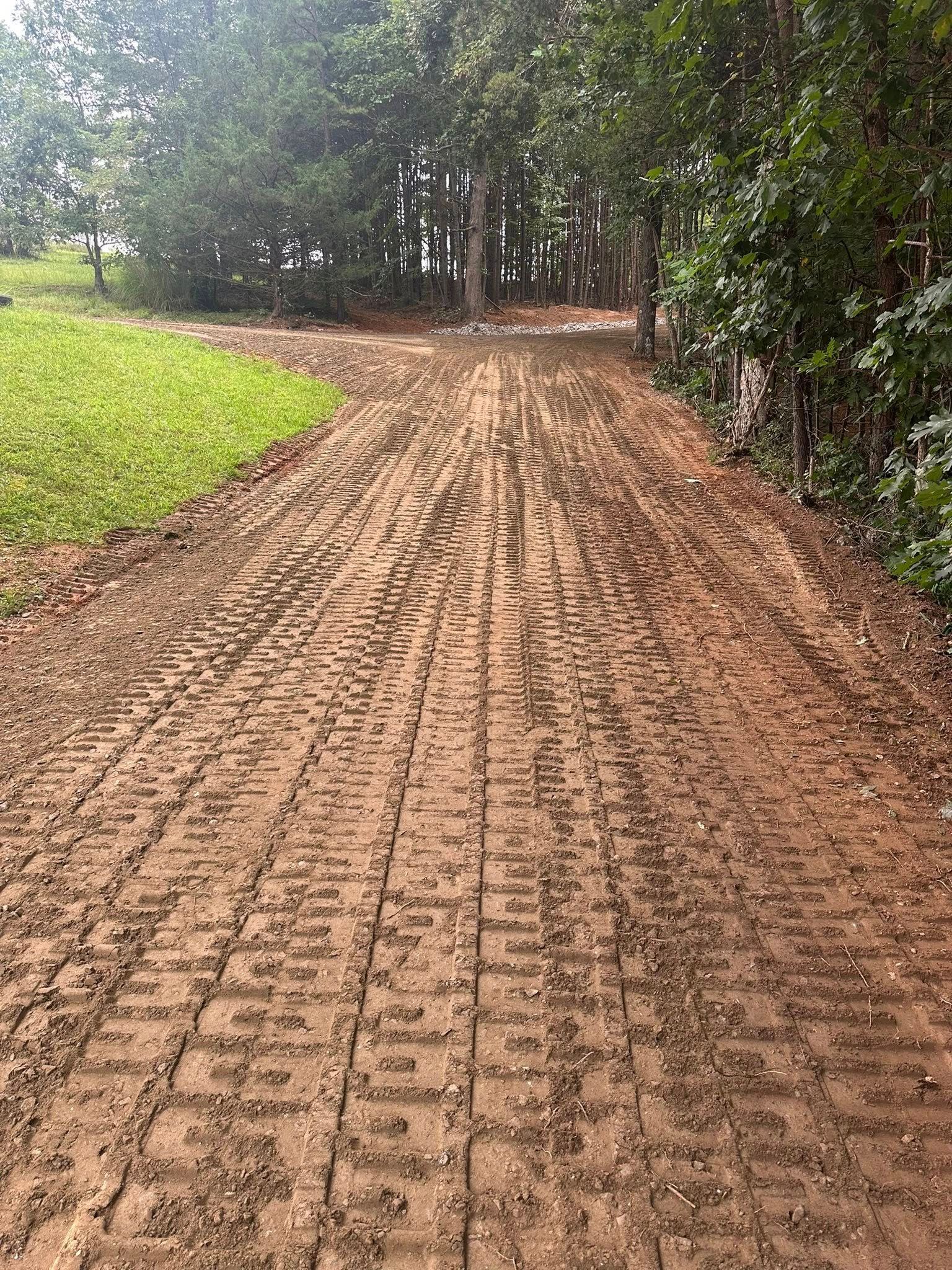 A muddy dirt path marked by deep tire tracks leads toward a line of trees in a wooded area.