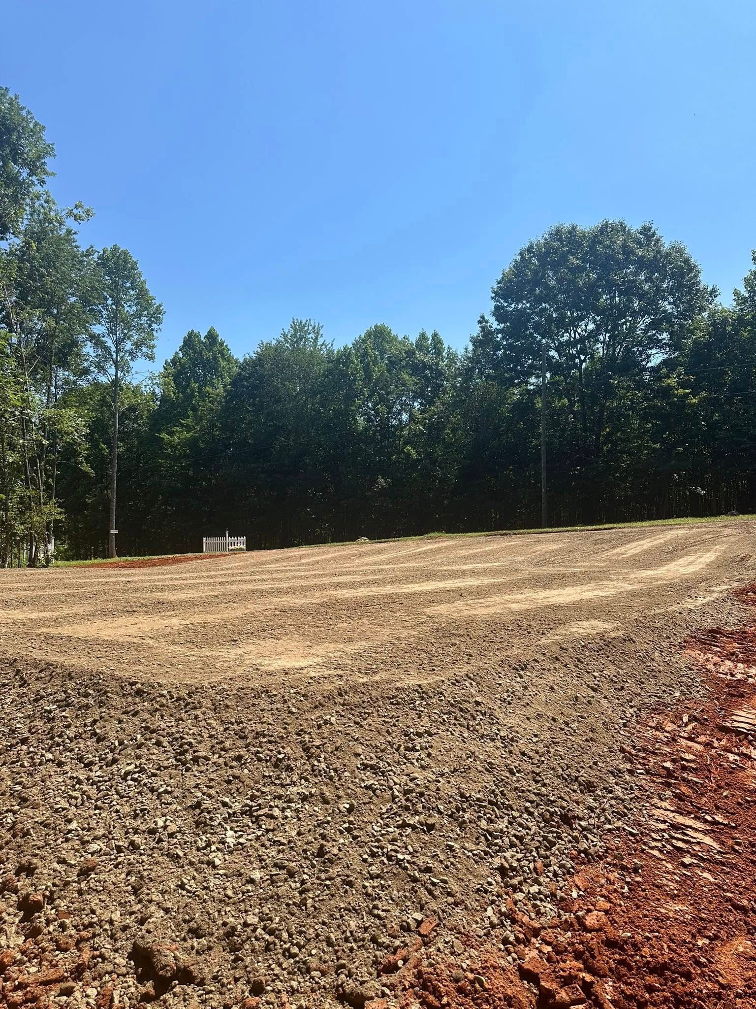 A cleared lot of land with a gravel base, bordered by a dense line of trees under a bright blue sky.