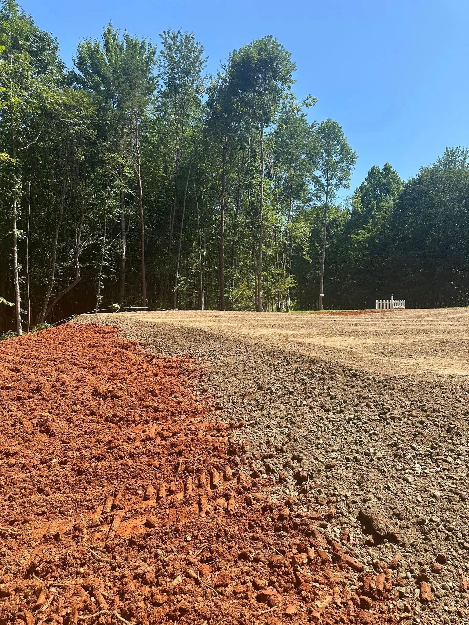A construction site featuring a foreground of reddish-brown clay next to gravel, with a dense forest line in the back.