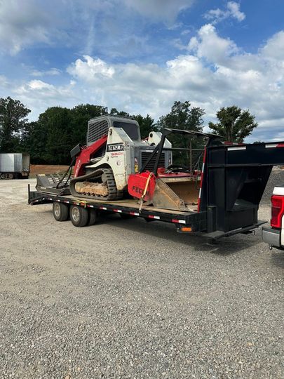 A white and red skid-steer loader sits on a flatbed trailer parked on a gravel lot under a cloudy sky.