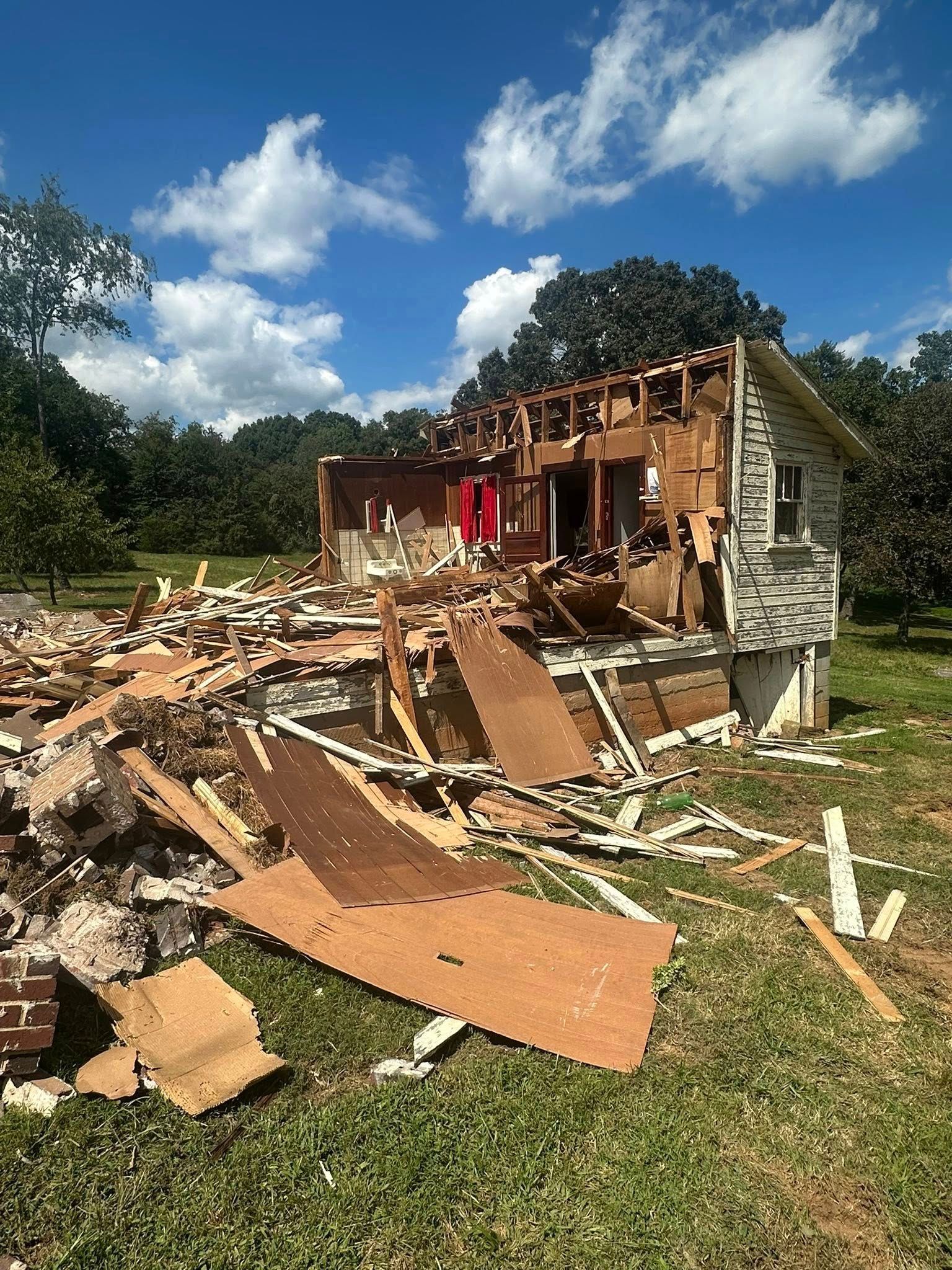 A house in ruins with collapsed walls and scattered wooden debris on a grassy lot under a sunny blue sky.