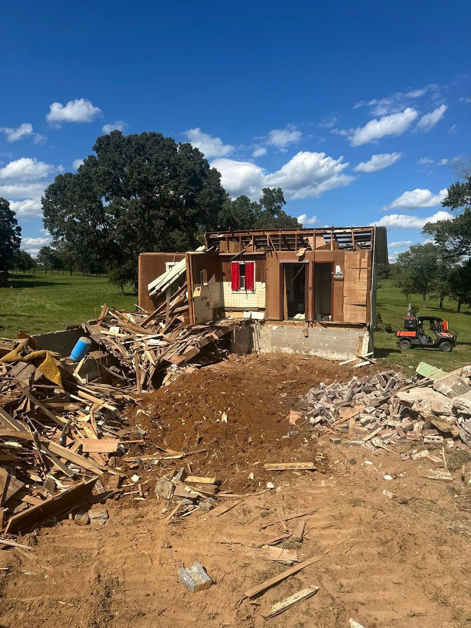 A partially demolished house stands in a rural, sunny landscape with a pile of building debris in the foreground.