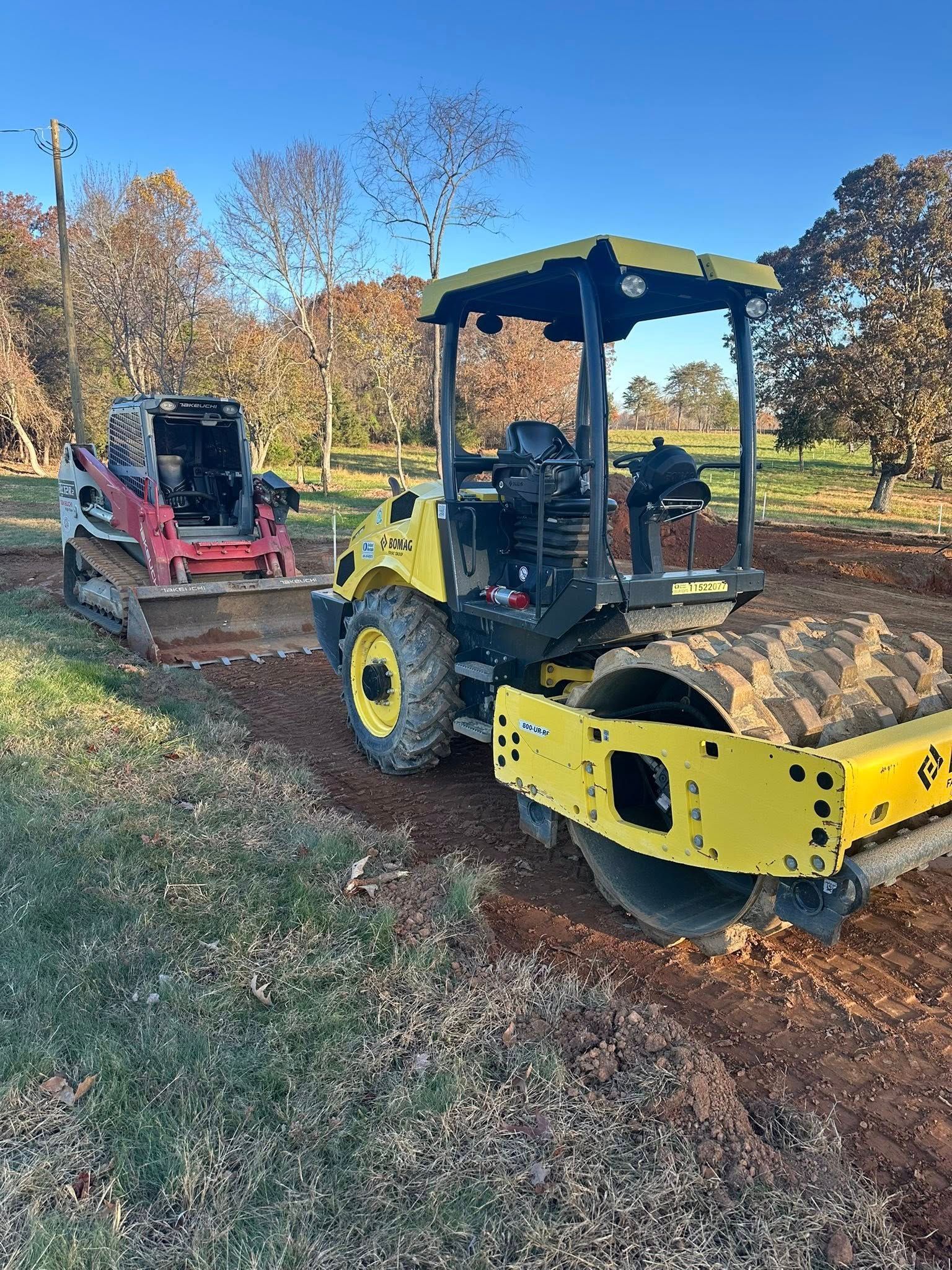 A yellow soil compactor and a red skid-steer loader parked on an earthen construction site under a clear blue sky.