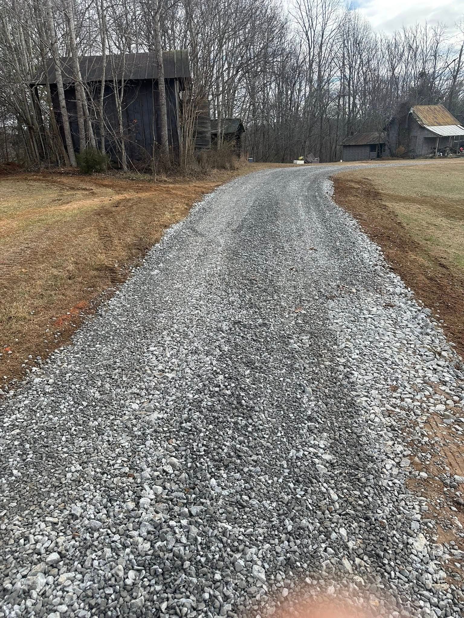 A gravel driveway leads toward a dark shed and a small house in a wooded, rural setting.