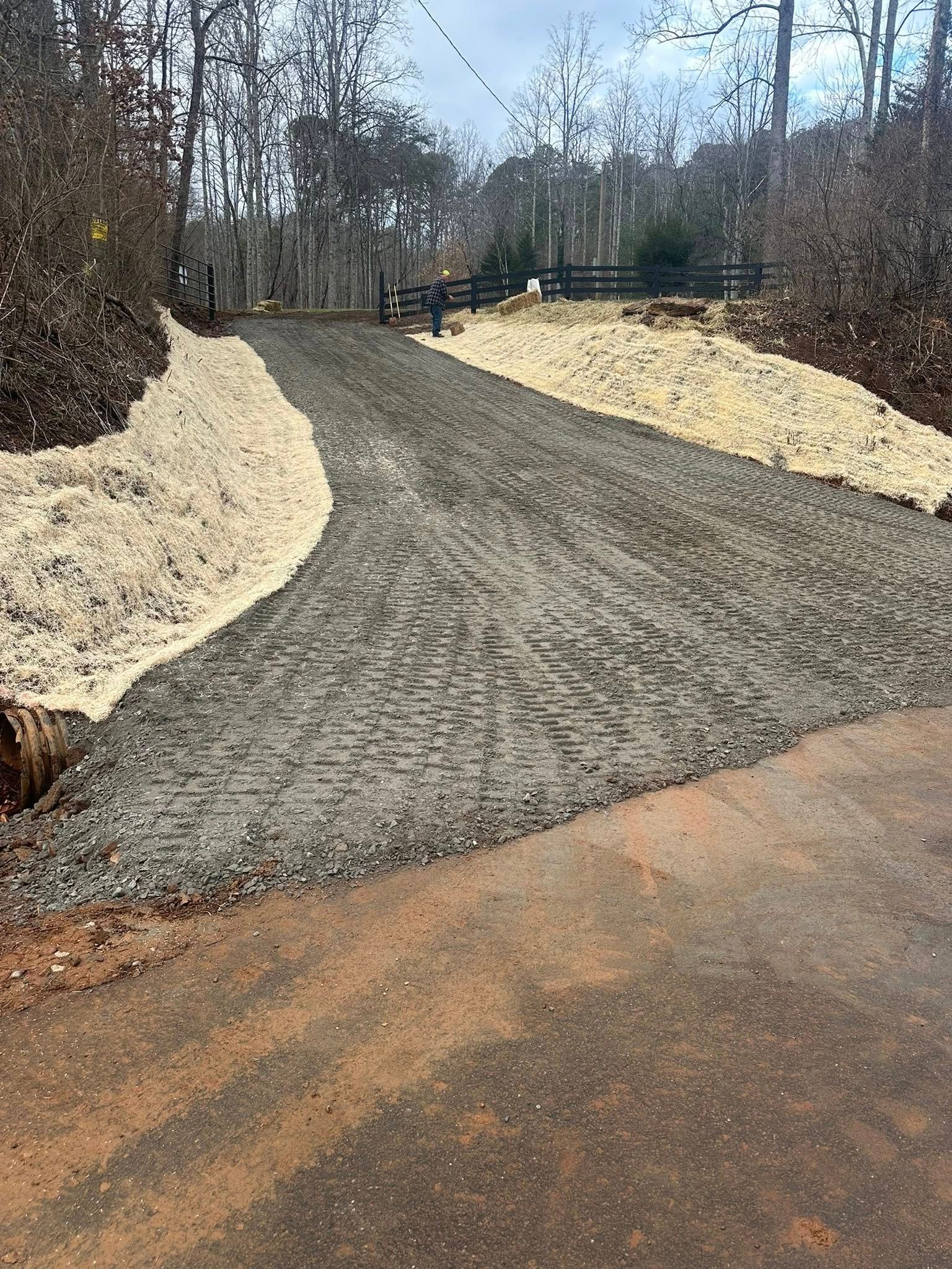 A gravel driveway entrance slopes upward between two banks covered in light-colored straw, leading toward a black fence.