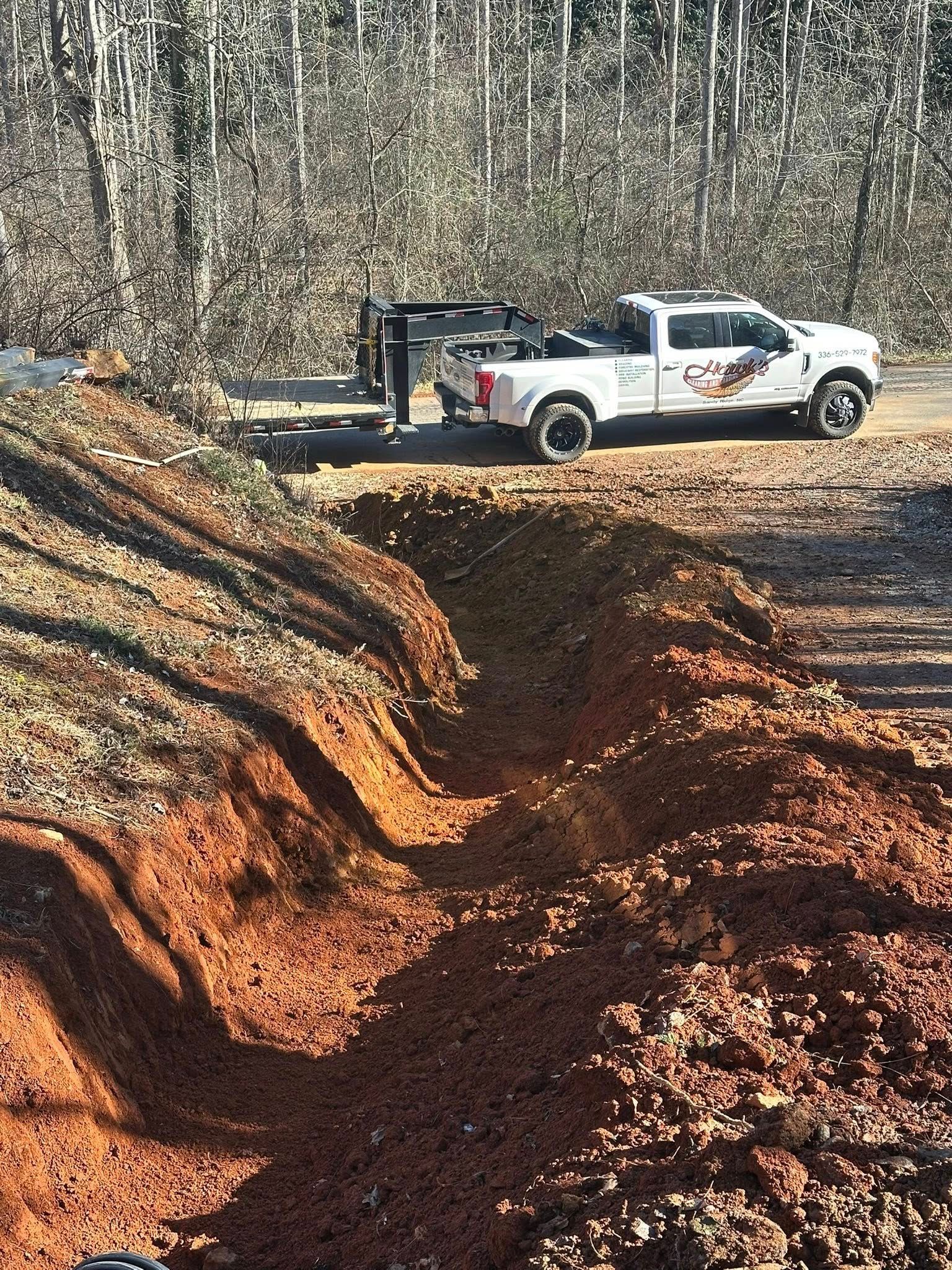 A white pickup truck with an attached flatbed trailer is parked on a dirt road near a freshly excavated trench.