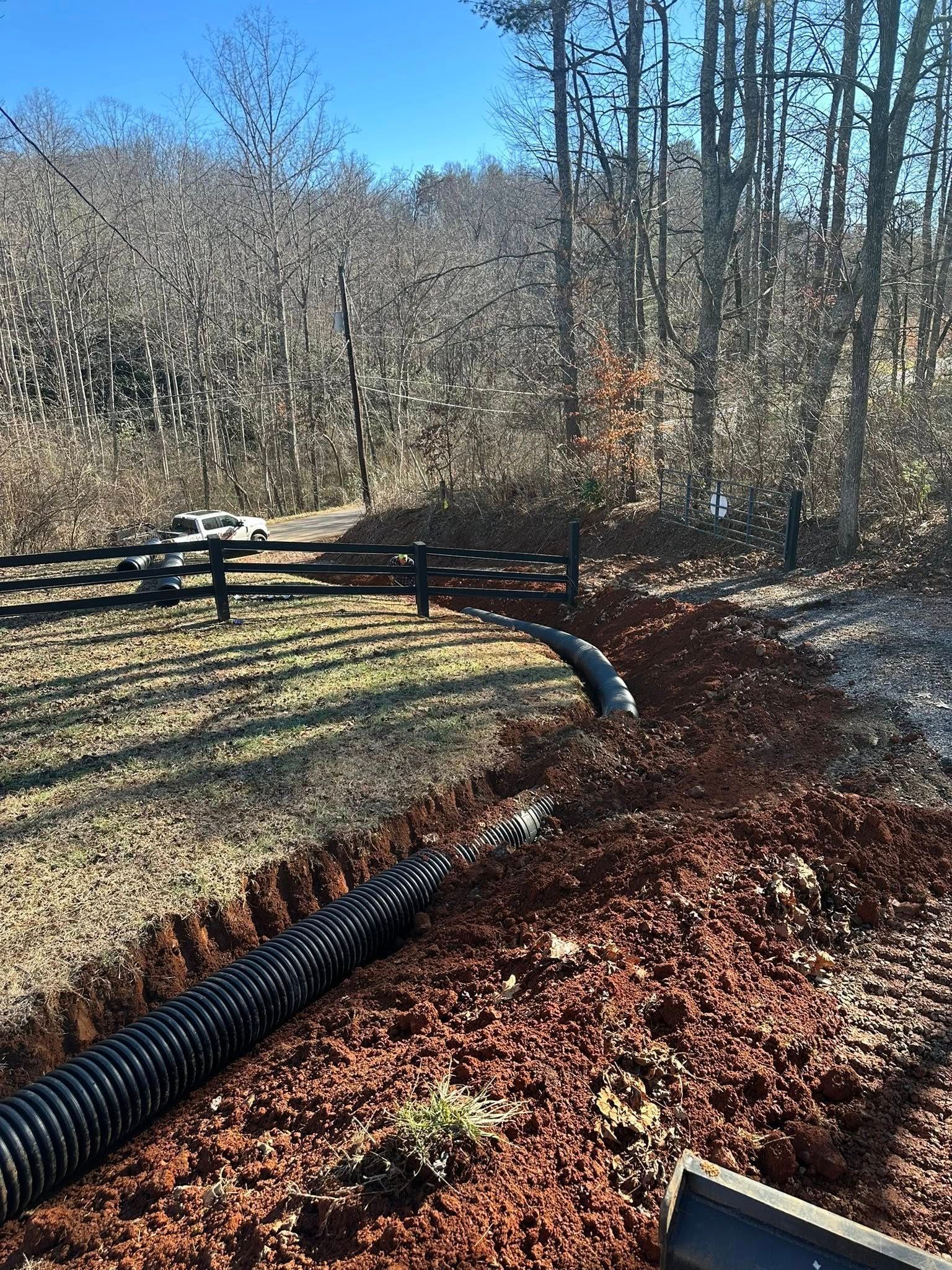 A black corrugated drainage pipe laid along a dirt trench beside a wooden fence in a wooded landscape.