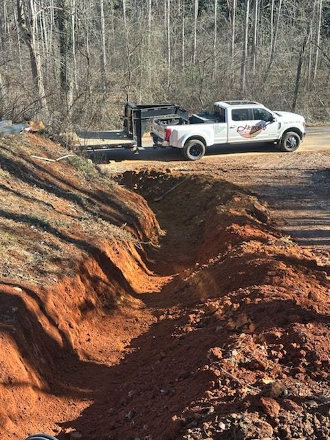 A white pickup truck with a trailer parked near a freshly excavated trench in a dirt field.