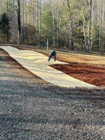 A person bends over while installing a light-colored erosion control mat on a patch of red earth near a forest.