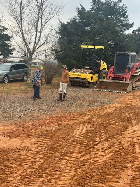 Two people stand talking on a gravel path near construction equipment, a yellow compactor, a red skid steer, and a car.