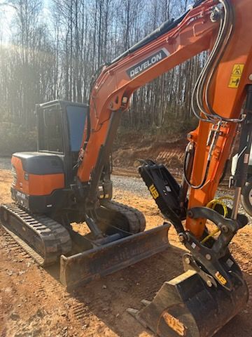 An orange and black DEVELON compact excavator parked on a dirt construction site against a backdrop of trees.