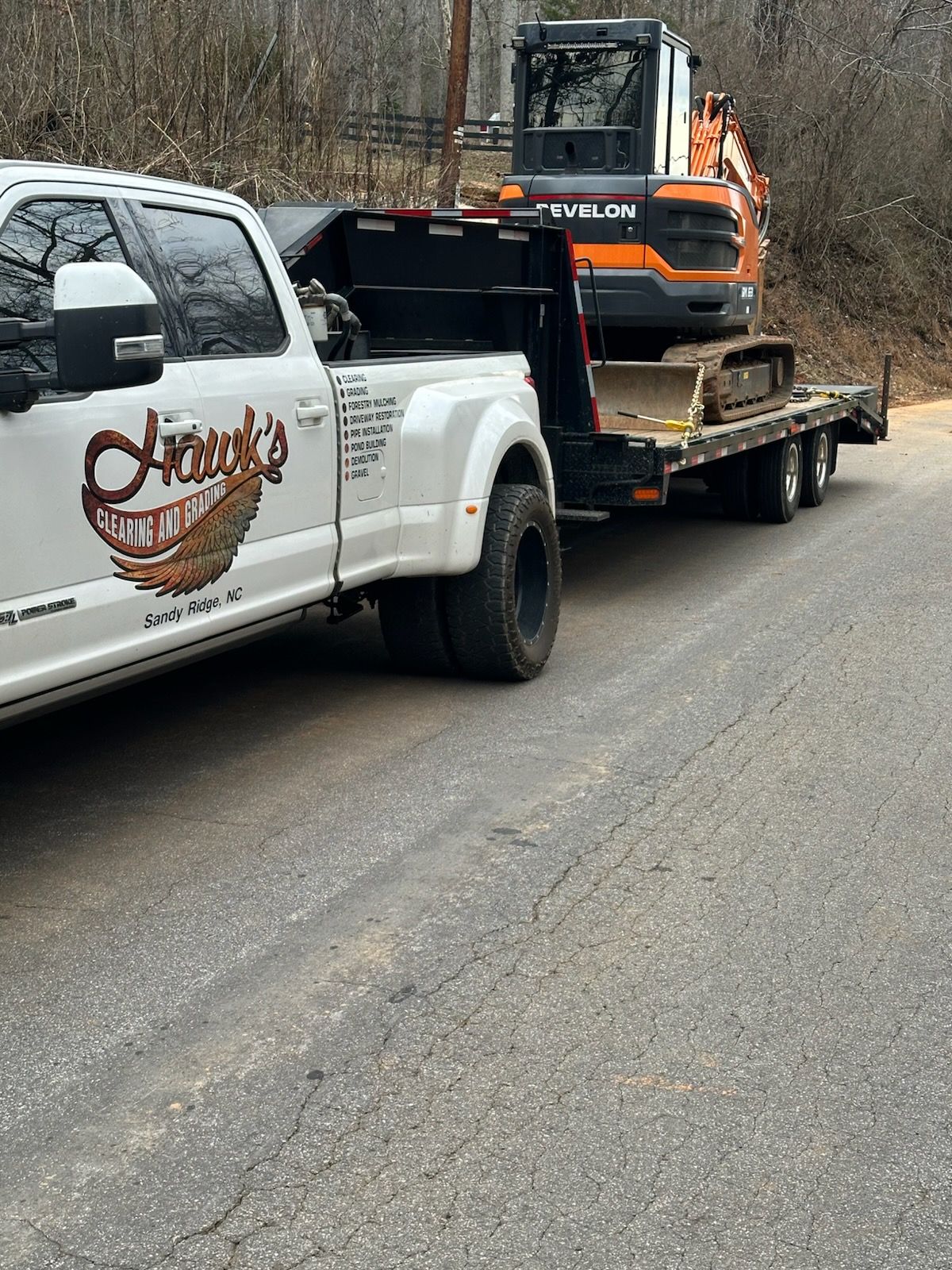 A white dual-wheel pickup truck hauling an orange excavator on a flatbed trailer on an asphalt road.
