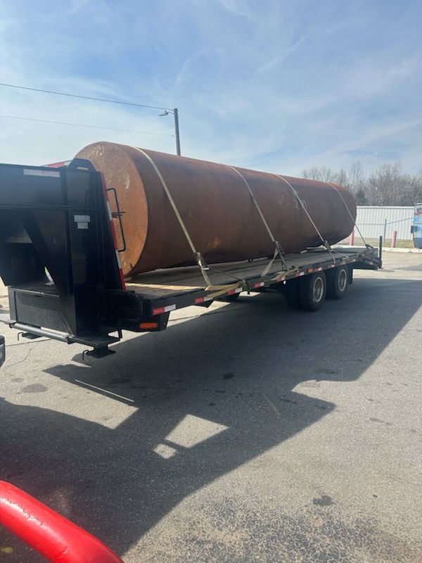 A flatbed trailer carrying a large, rusted cylindrical steel tank on an asphalt lot under a blue sky.