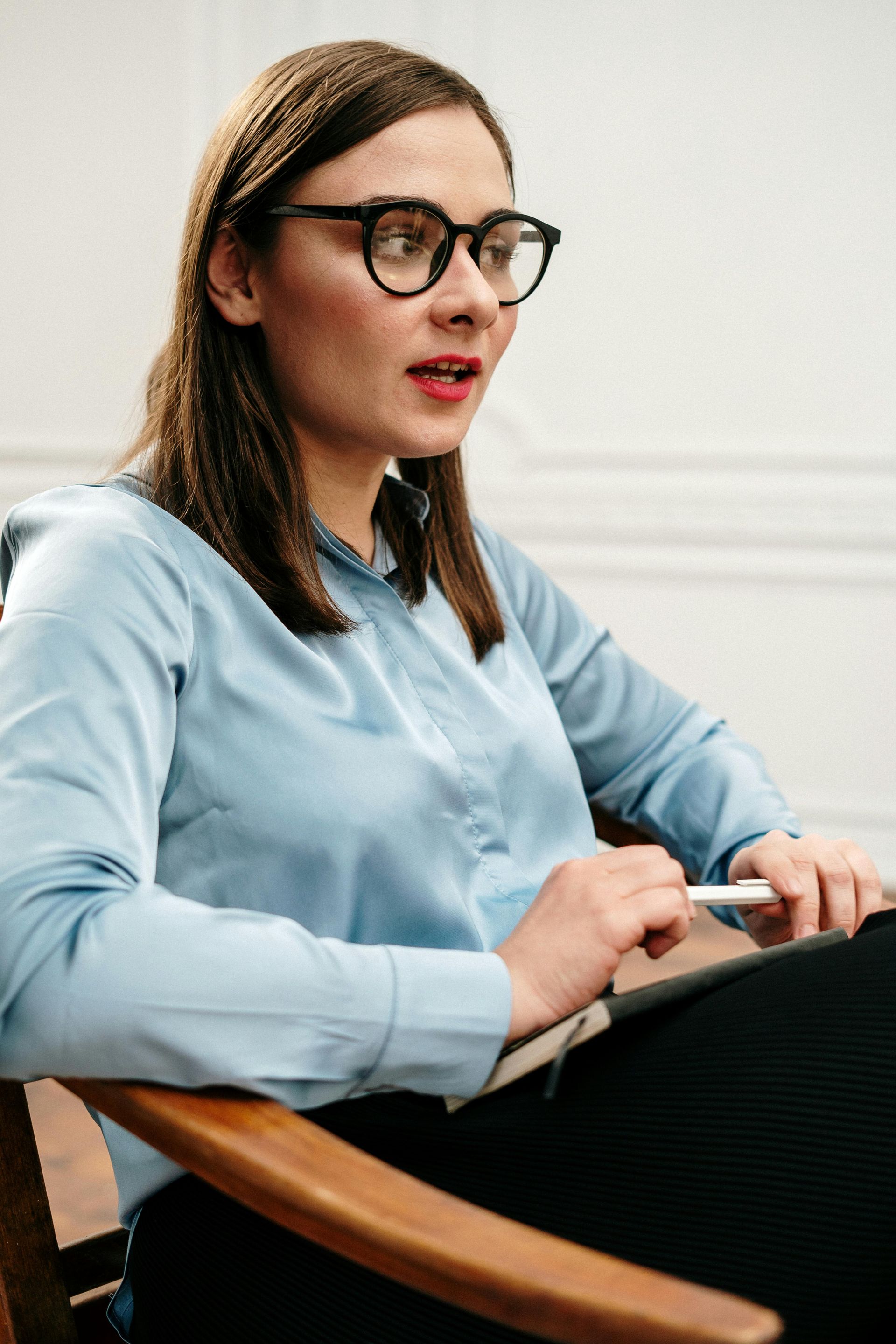 Woman in glasses speaking while seated in a chair, wearing a light blue shirt and holding a tablet.