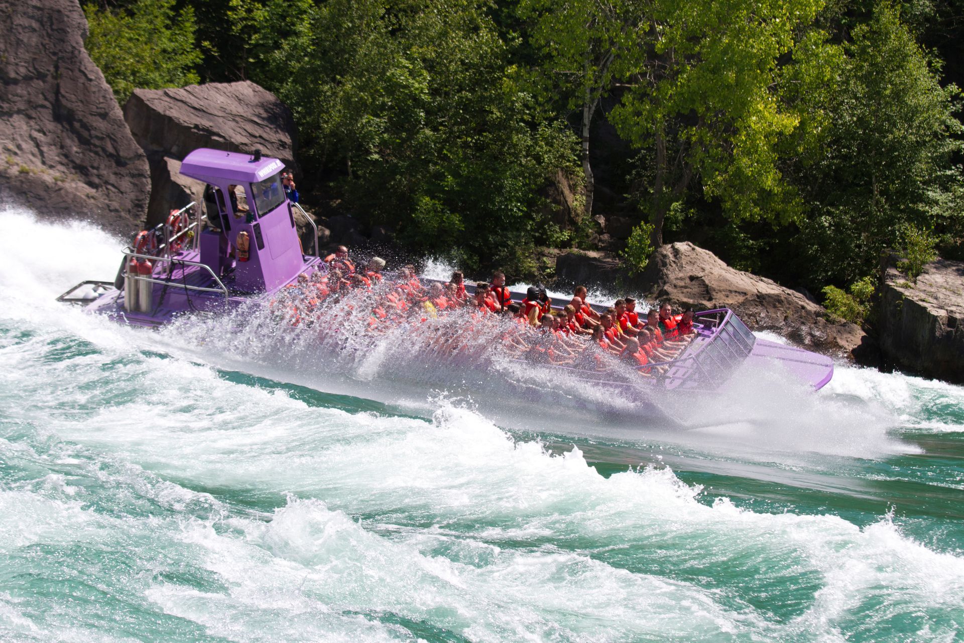 A purple Whirlpool Jet Boat speeds through whitewater rapids as spray rises beside its passengers.