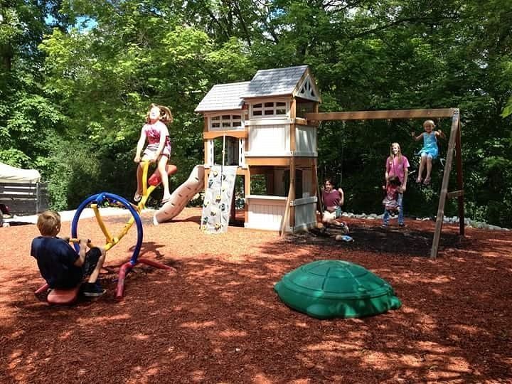 Children playing on a backyard playground with a slide, swing, and climbing toys under trees.