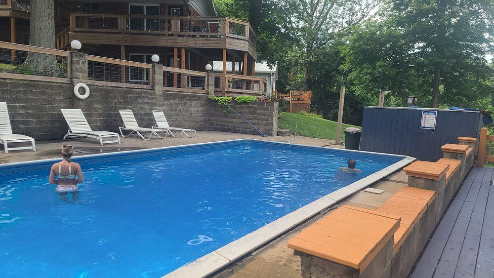 Outdoor pool with a swimmer, lounge chairs, and a wooden deck beside a wooded hillside resort