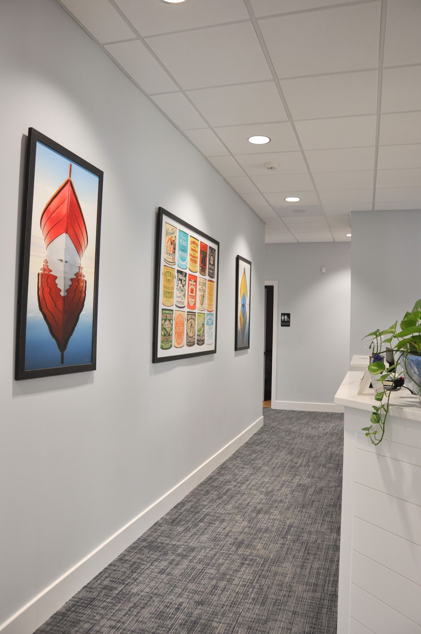 Hallway with artwork on the walls, a gray carpet, and a white reception desk with plants.
