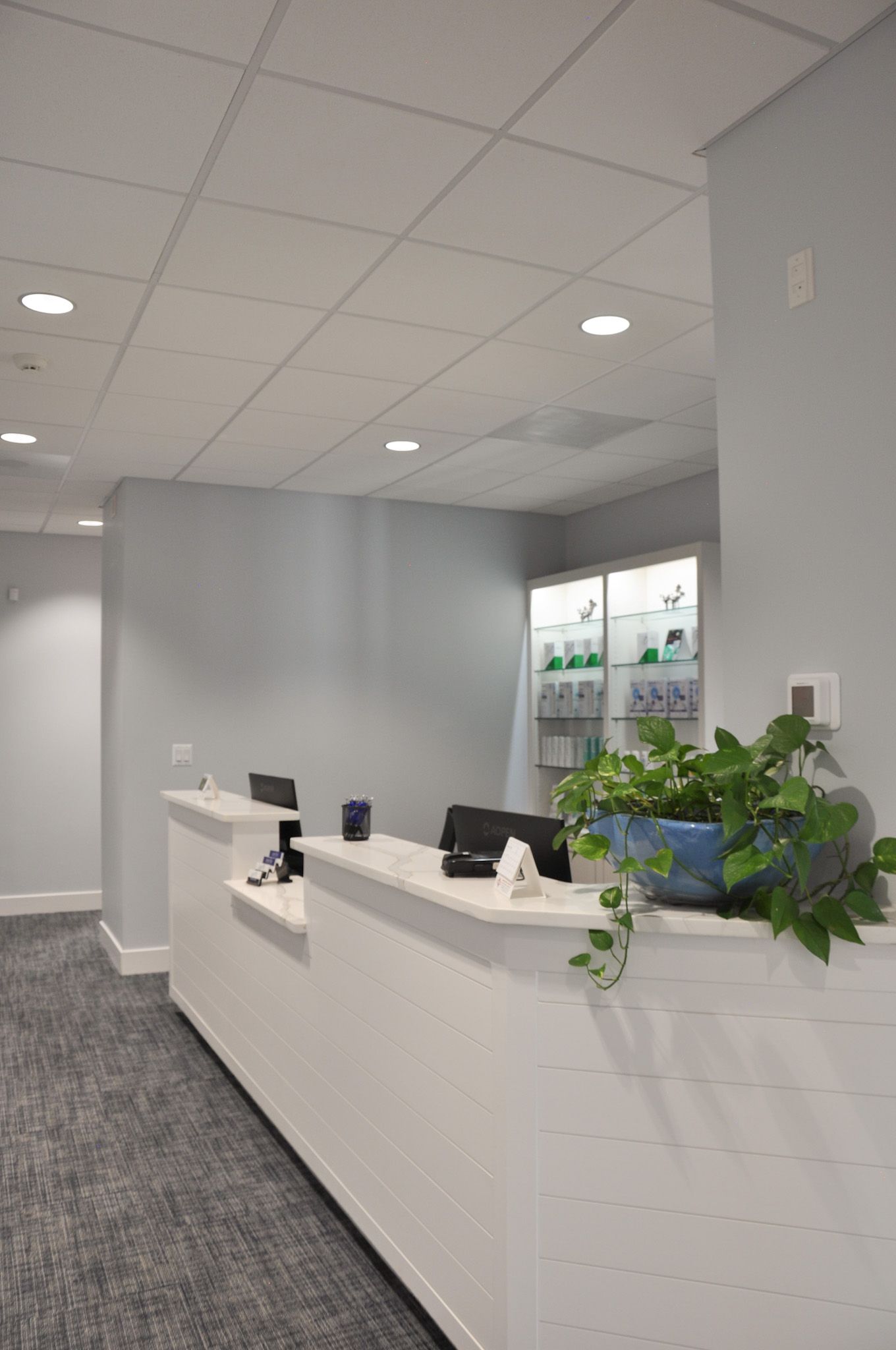 White reception desk in a medical office, with a computer, plants, and product display. Light blue walls and carpet.