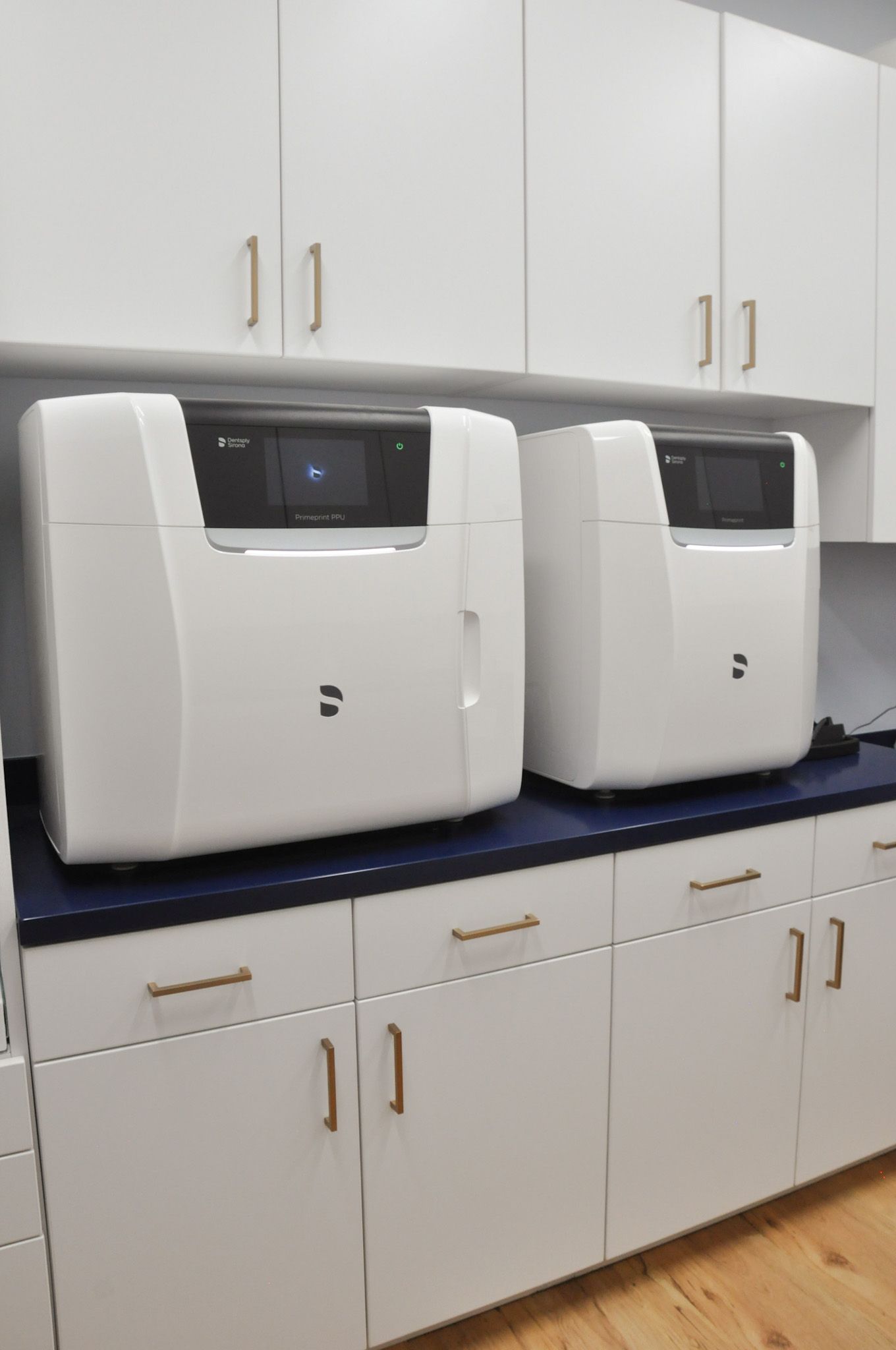 Two white dental machines on a counter with white cabinets and gold handles.